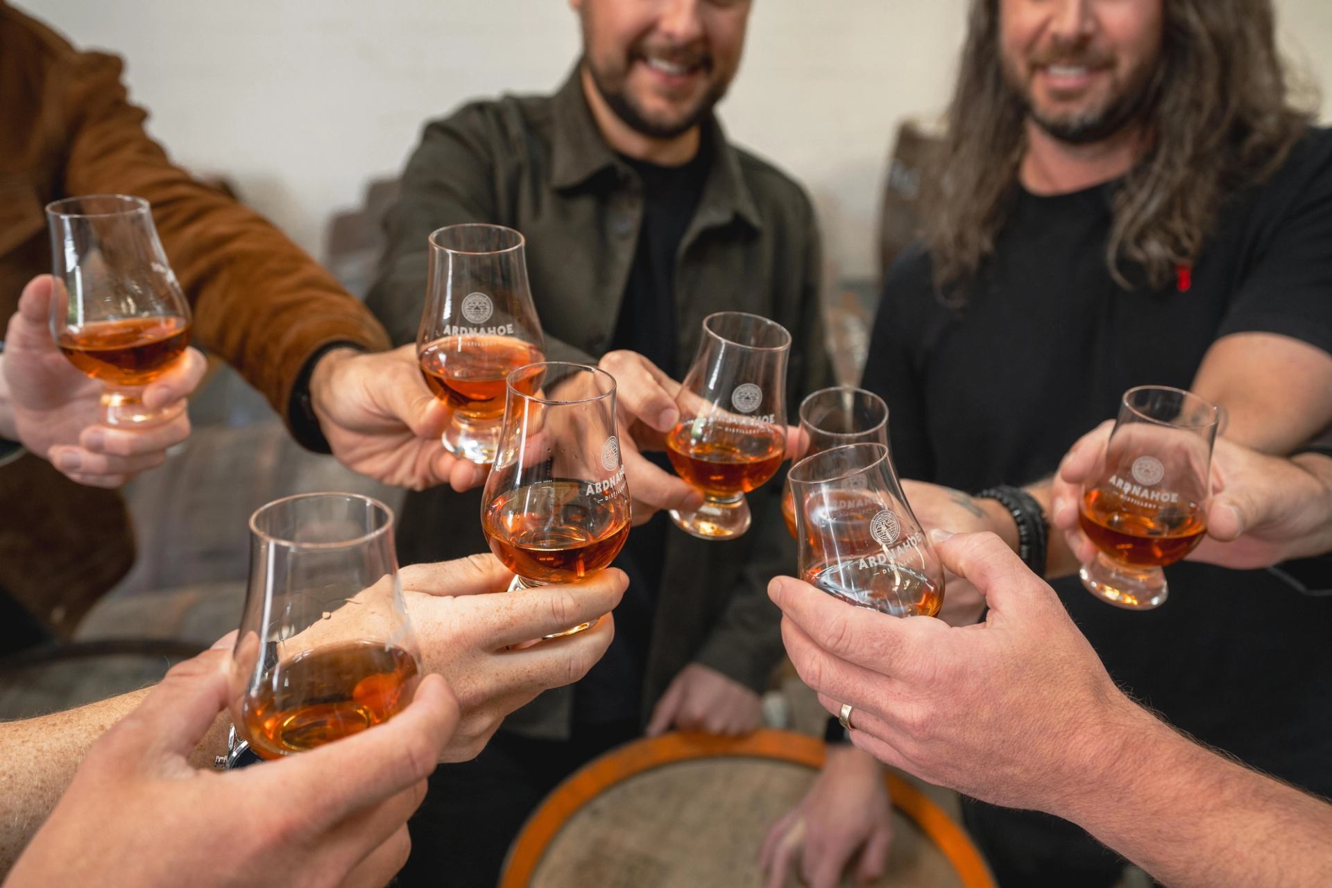 Hands holding whisky glasses together in a toast above a wooden barrel.