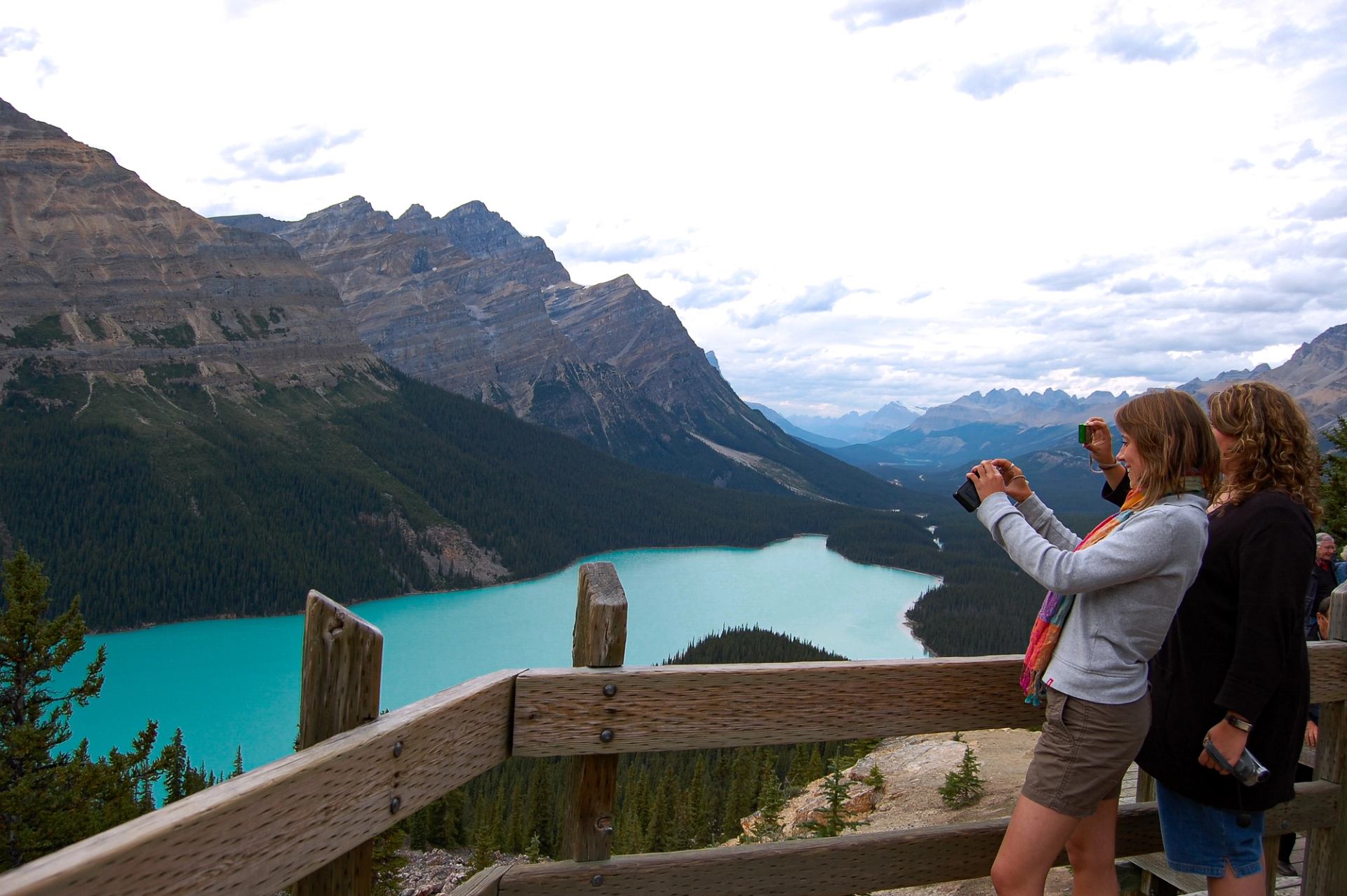 People taking photos of a turquoise lake surrounded by dramatic mountain peaks.