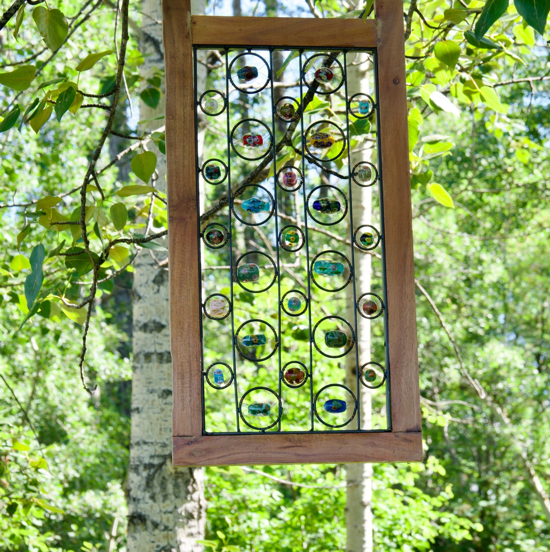 Wooden-framed glass art panel with colourful circles hanging among leafy green trees.