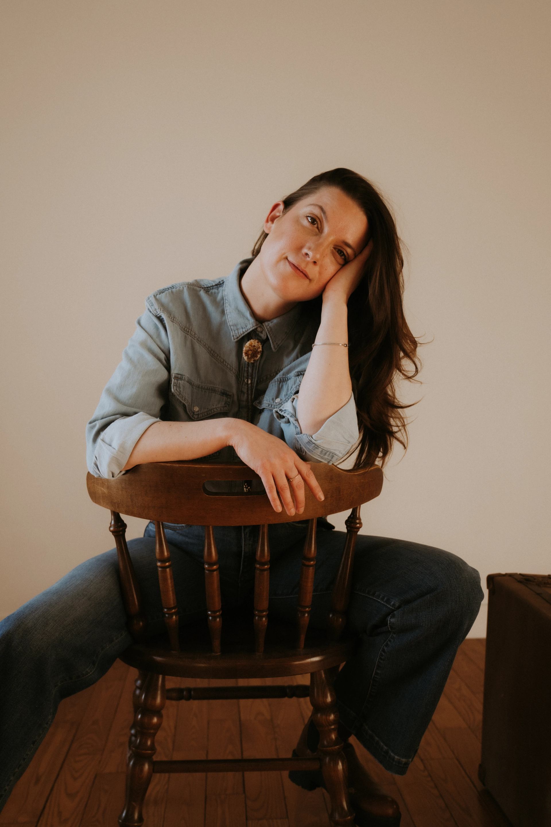 Person in denim shirt sits backwards on wooden chair, head resting on hand.