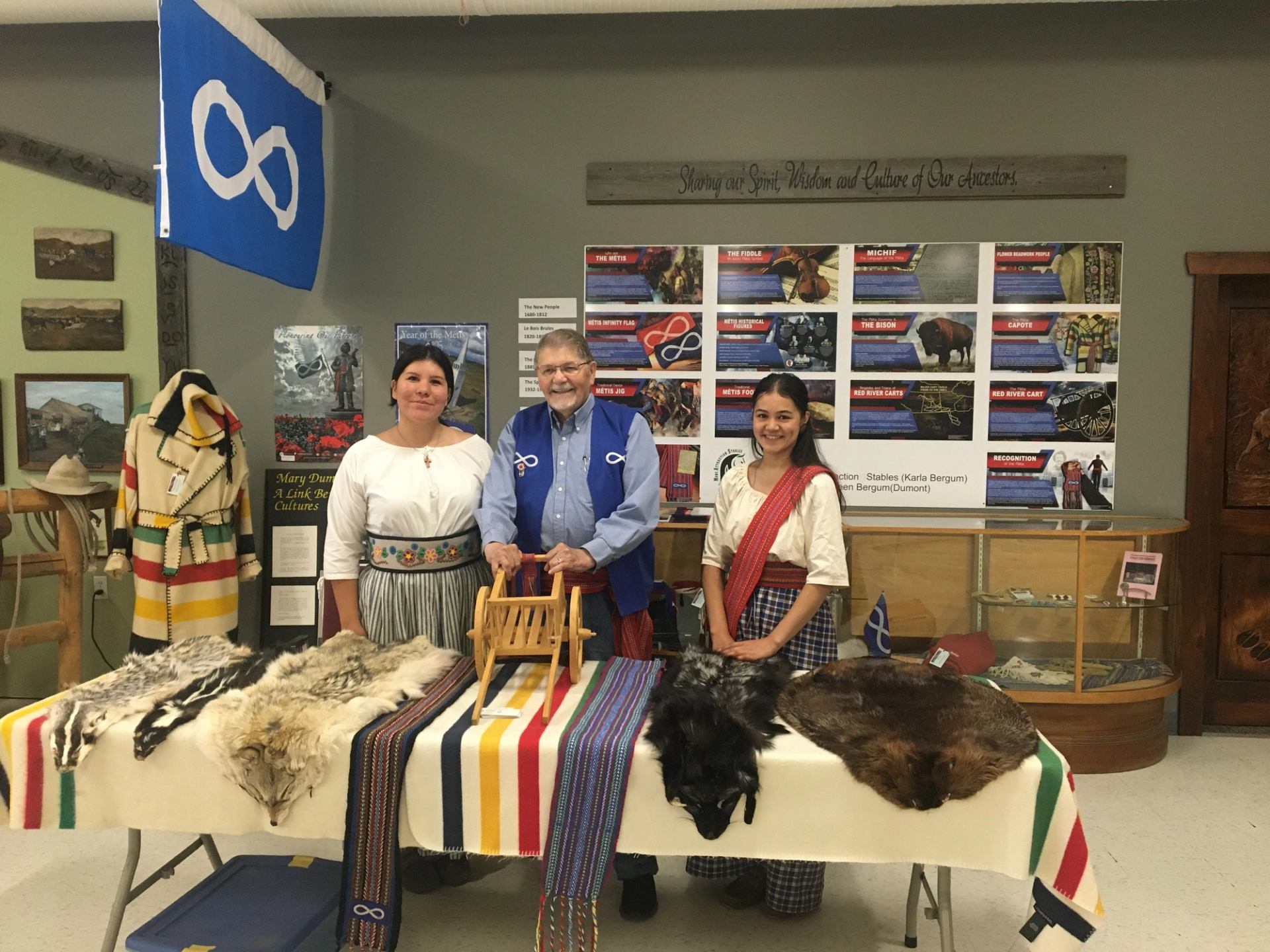 Three smiling presenters stand behind a display table showcasing Métis sashes, traditional animal furs, and a miniature Red River cart. The background features educational poster boards and a large blue Métis flag hanging prominently on the wall.