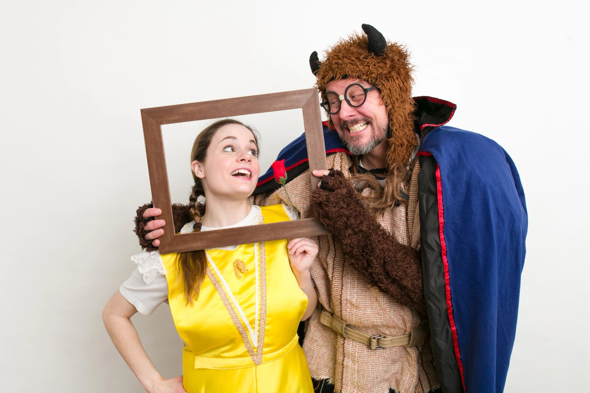 Two people in fairy-tale costumes hold an empty frame and a red rose.