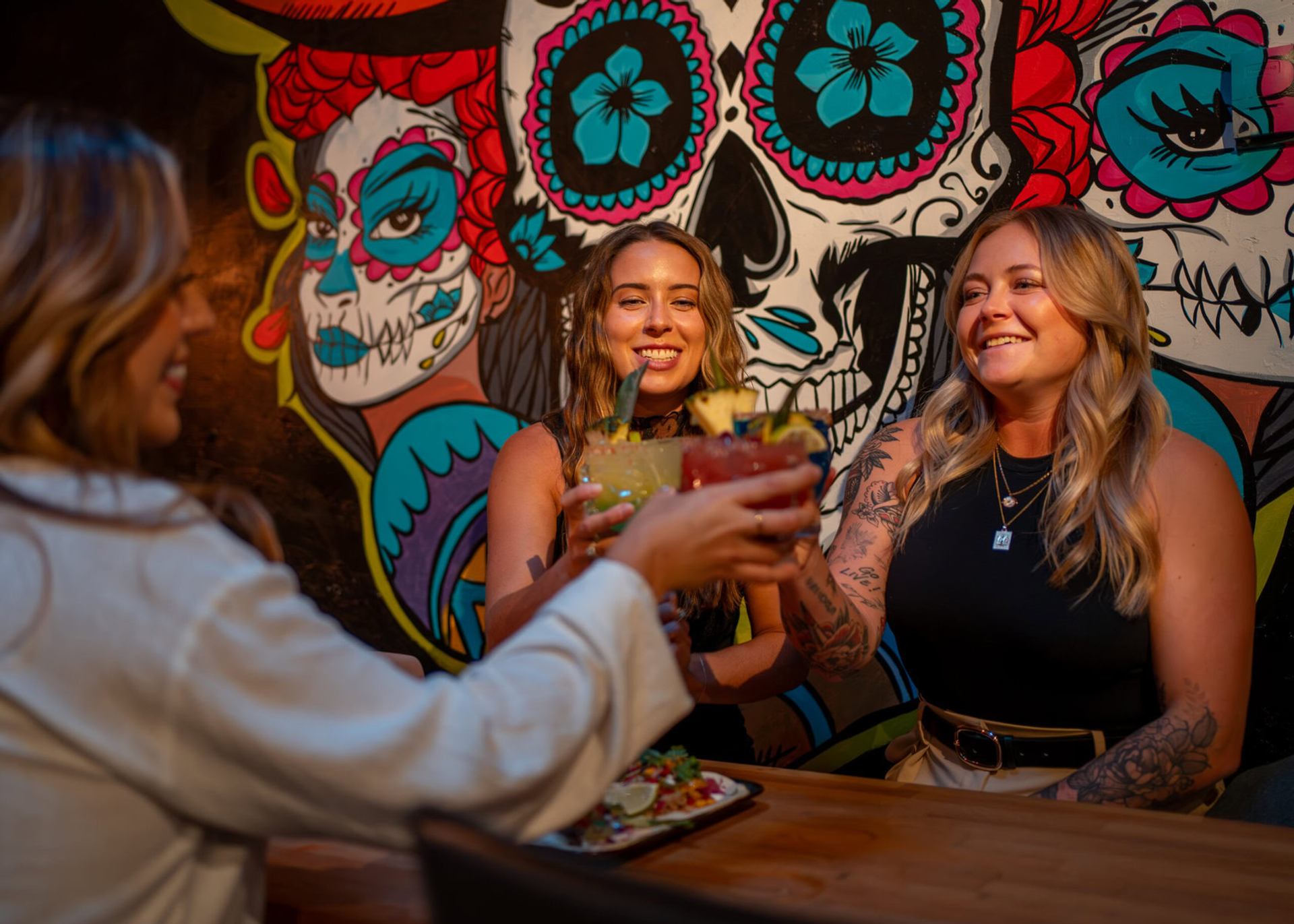 Friends raise colourful cocktails at a bar table before a vibrant skull mural.