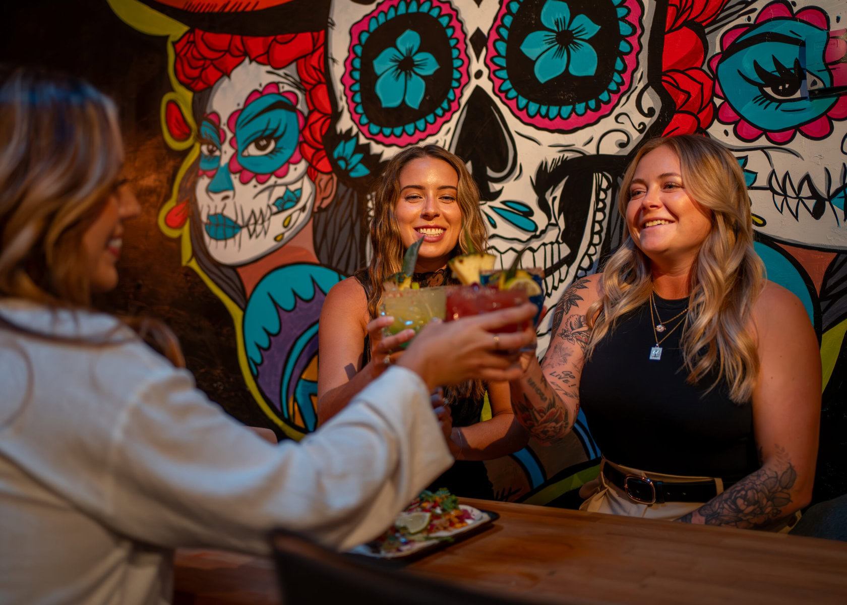 Friends raise colourful cocktails at a bar table before a vibrant skull mural.
