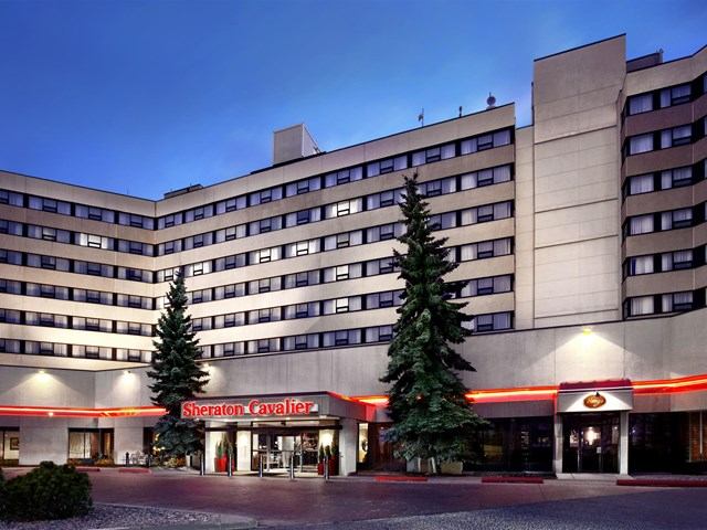 Exterior of Sheraton Cavalier Calgary Hotel with red signage and evergreen trees.