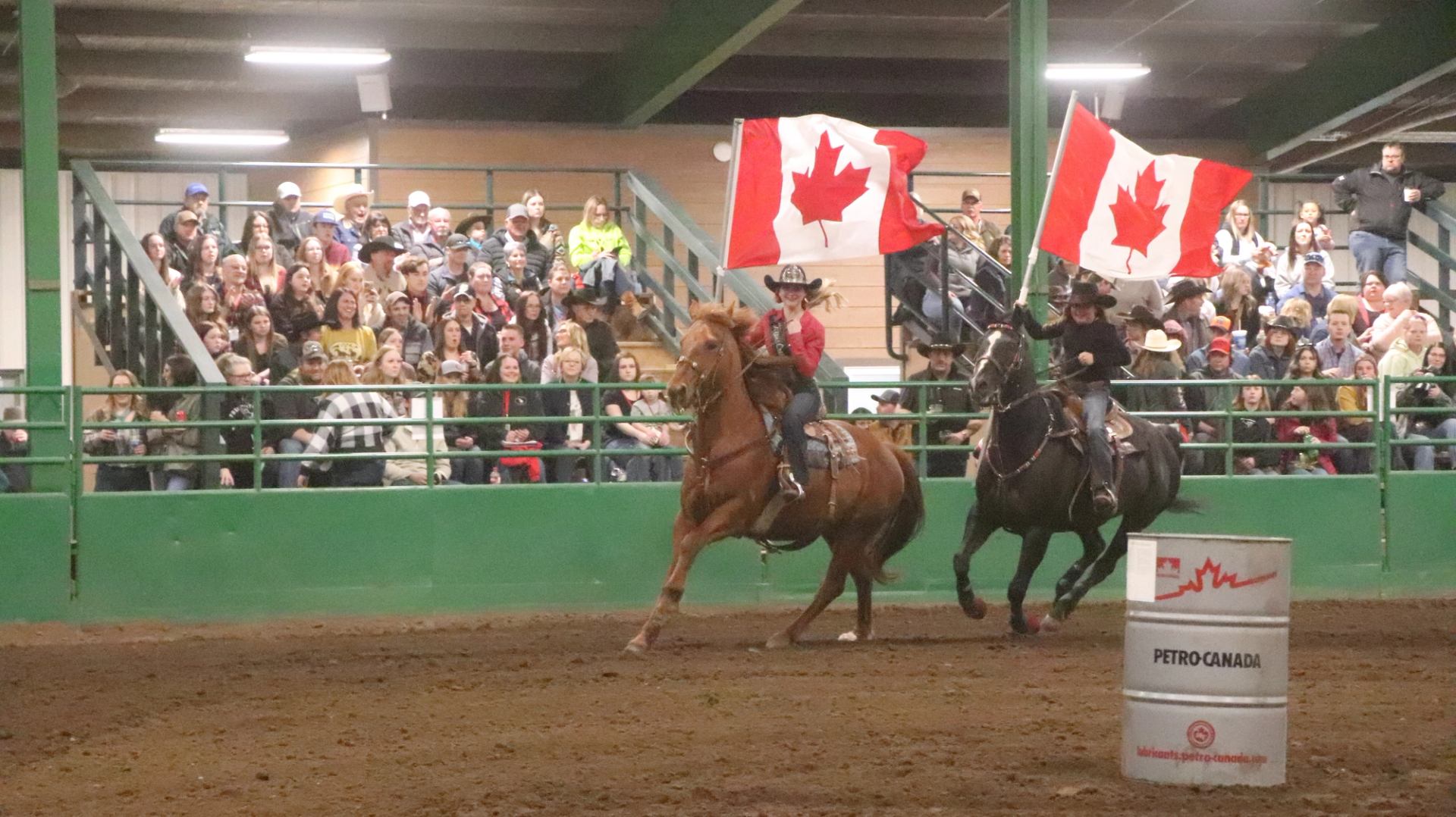 Riders on horses carry Canadian flags in indoor arena before large seated crowd.