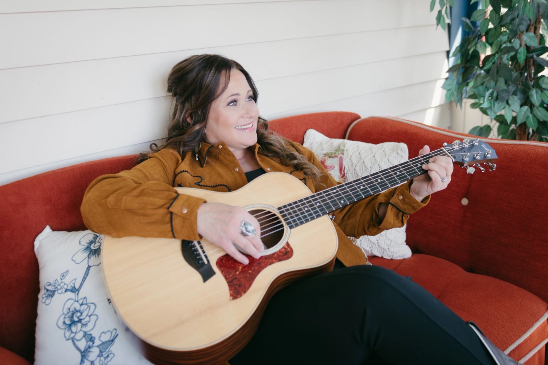 Person reclining on red couch, playing an acoustic guitar near indoor plant.