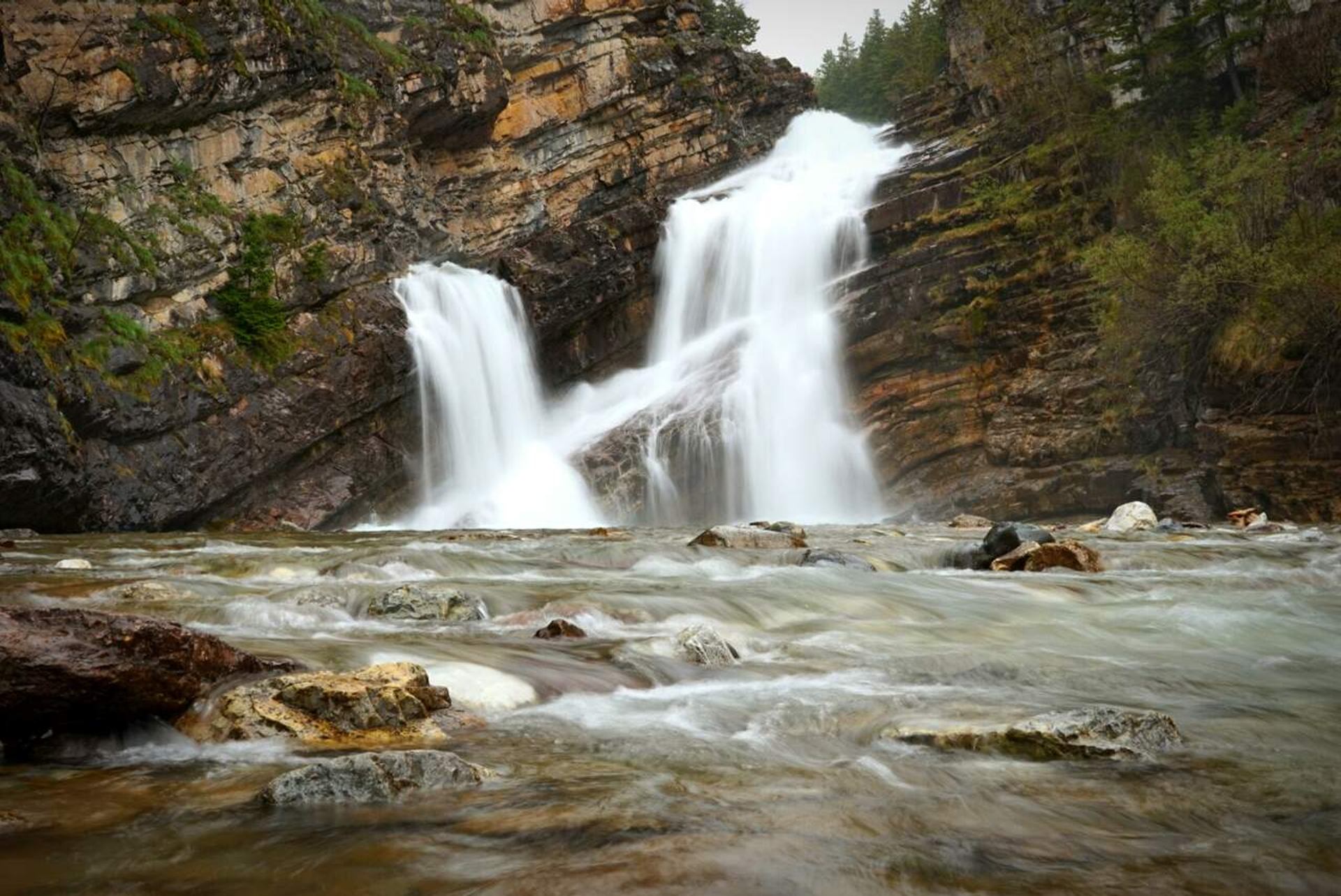 Twin waterfall cascades over layered rock cliffs into a shallow, fast-flowing river.