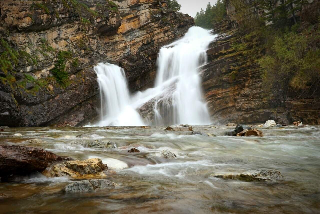 Twin waterfall cascades over layered rock cliffs into a shallow, fast-flowing river.