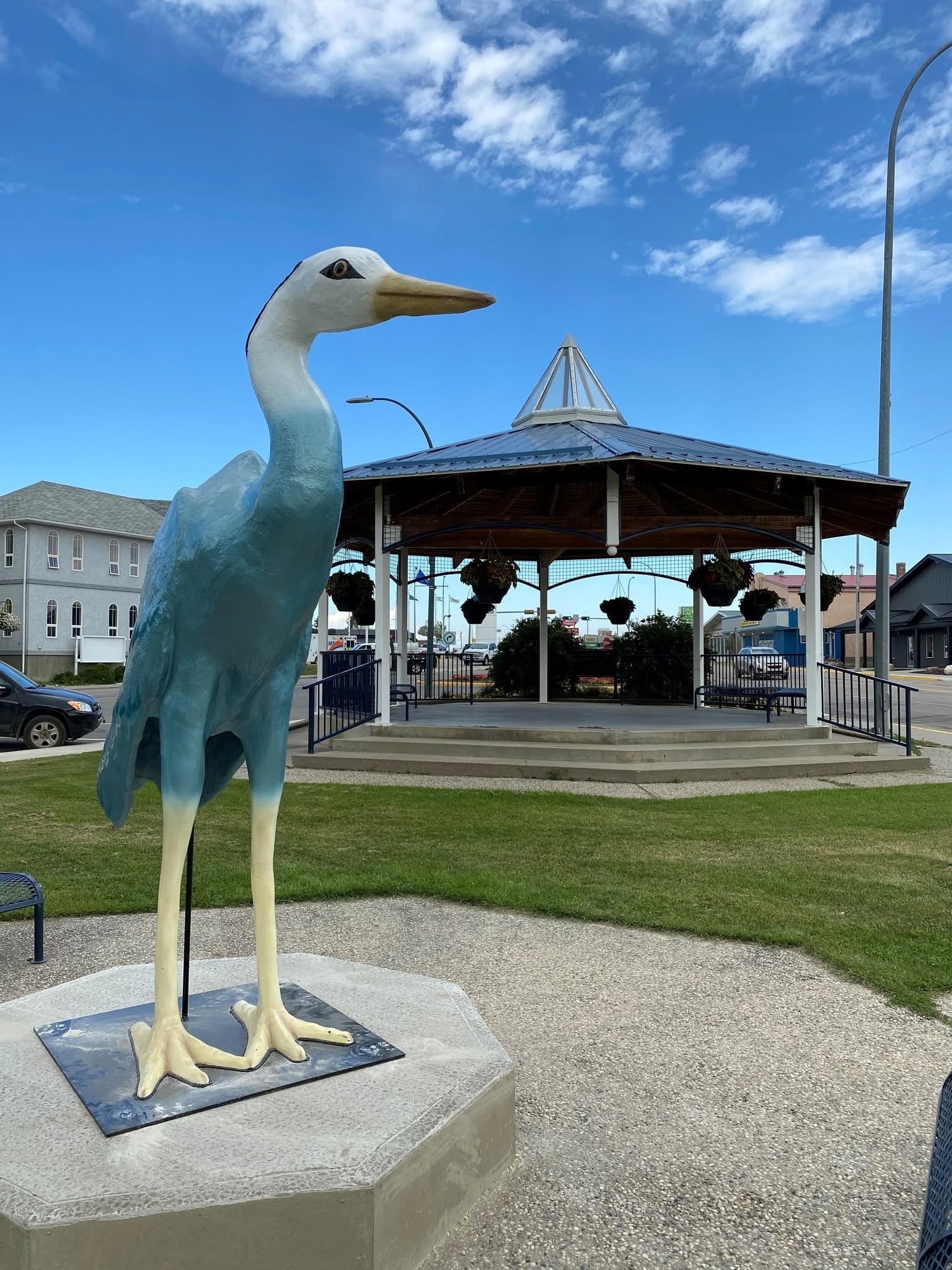 A large blue heron statue stands on a raised concrete pedestal in a grassy public area. The bird sculpture has a long neck, pale blue body, cream-colored legs, and a yellow beak. Behind the statue, a covered gazebo with hanging flower baskets sits under a bright blue sky with scattered white clouds. Nearby buildings, parked vehicles, and streetlights are visible in the background.
