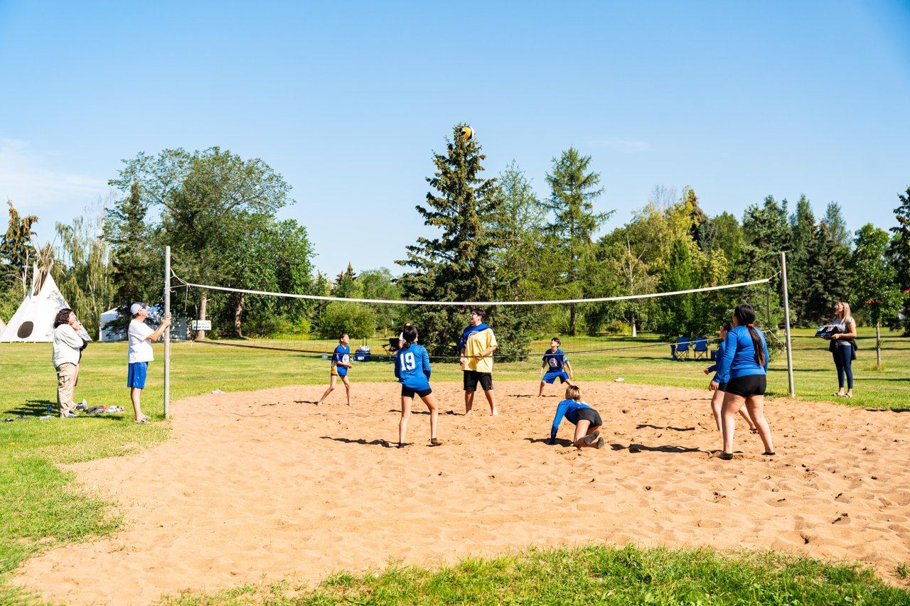 Youth volleyball players on a sandy court in a park with trees and teepee.