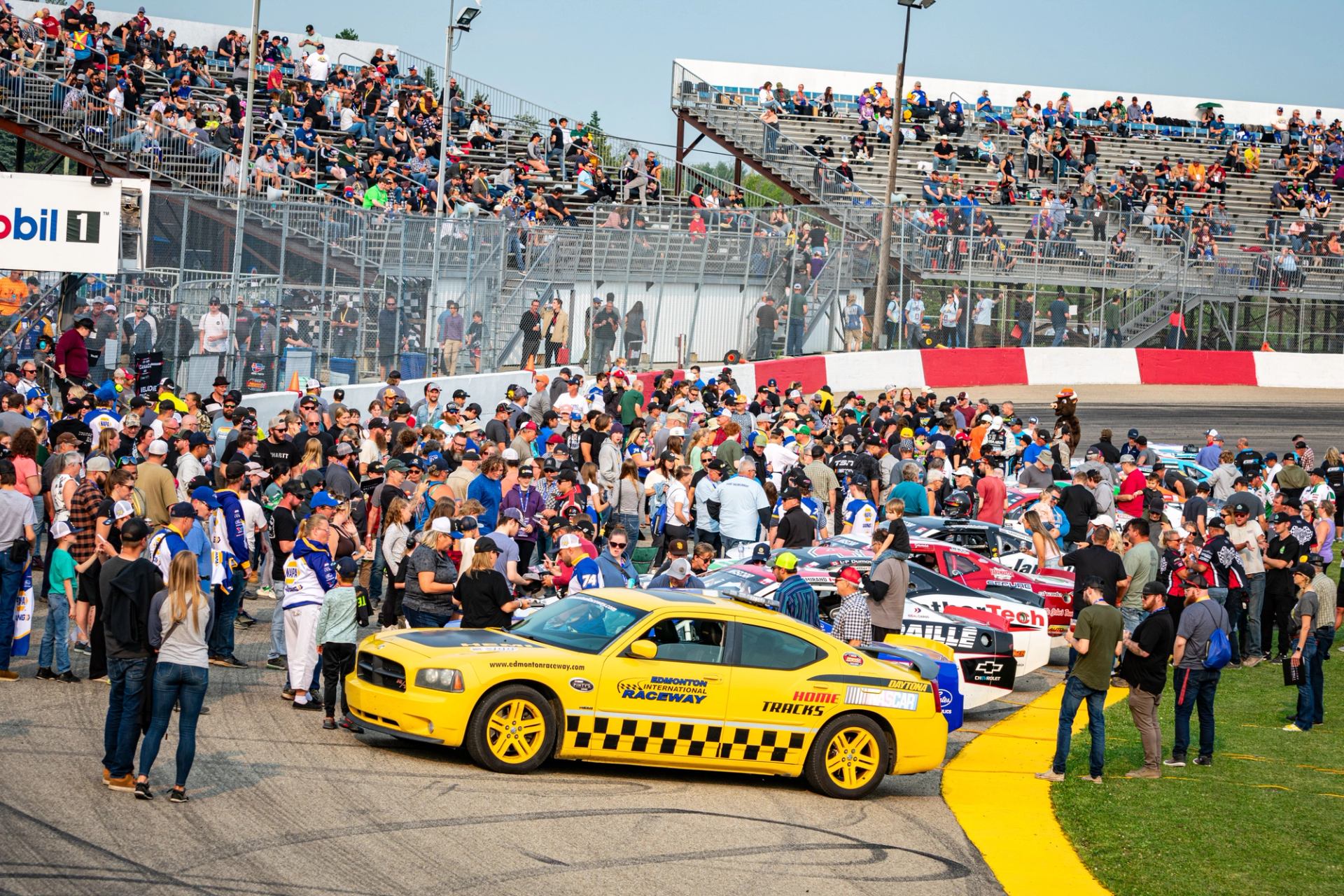 Large crowd surrounds NASCAR cars and a yellow pace car before the race begins.