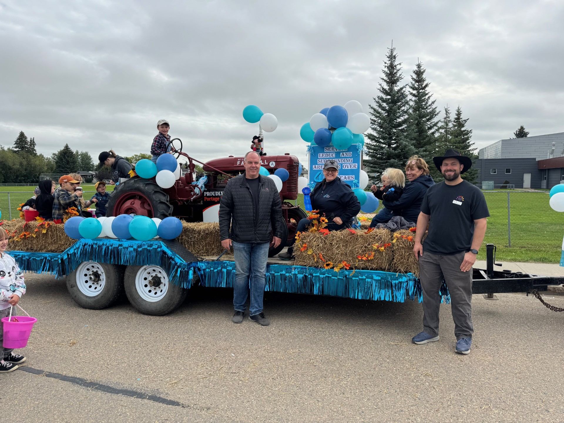 Parade float with tractor, hay bales, balloons and people posing on decorated trailer.
