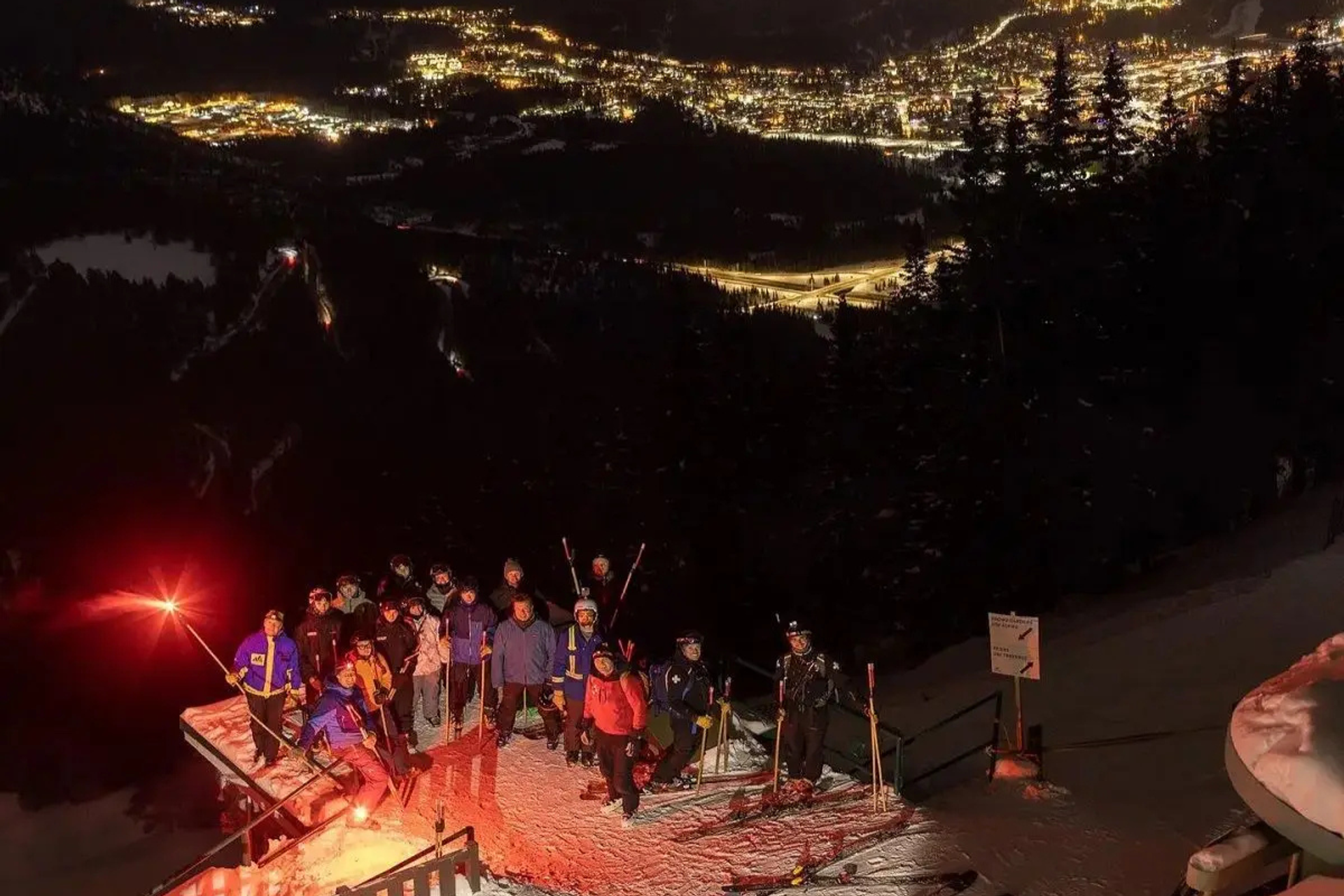Group with glowing torches stands on snowy platform overlooking a brightly lit mountain town.