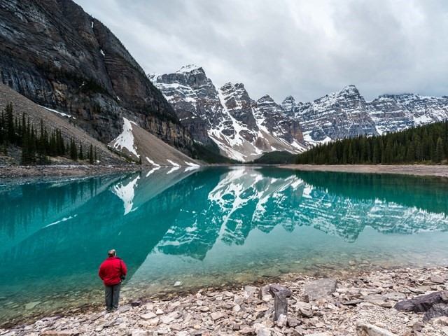 Person in a red jacket standing by a clear turquoise lake with snow-capped mountains reflected in the water.