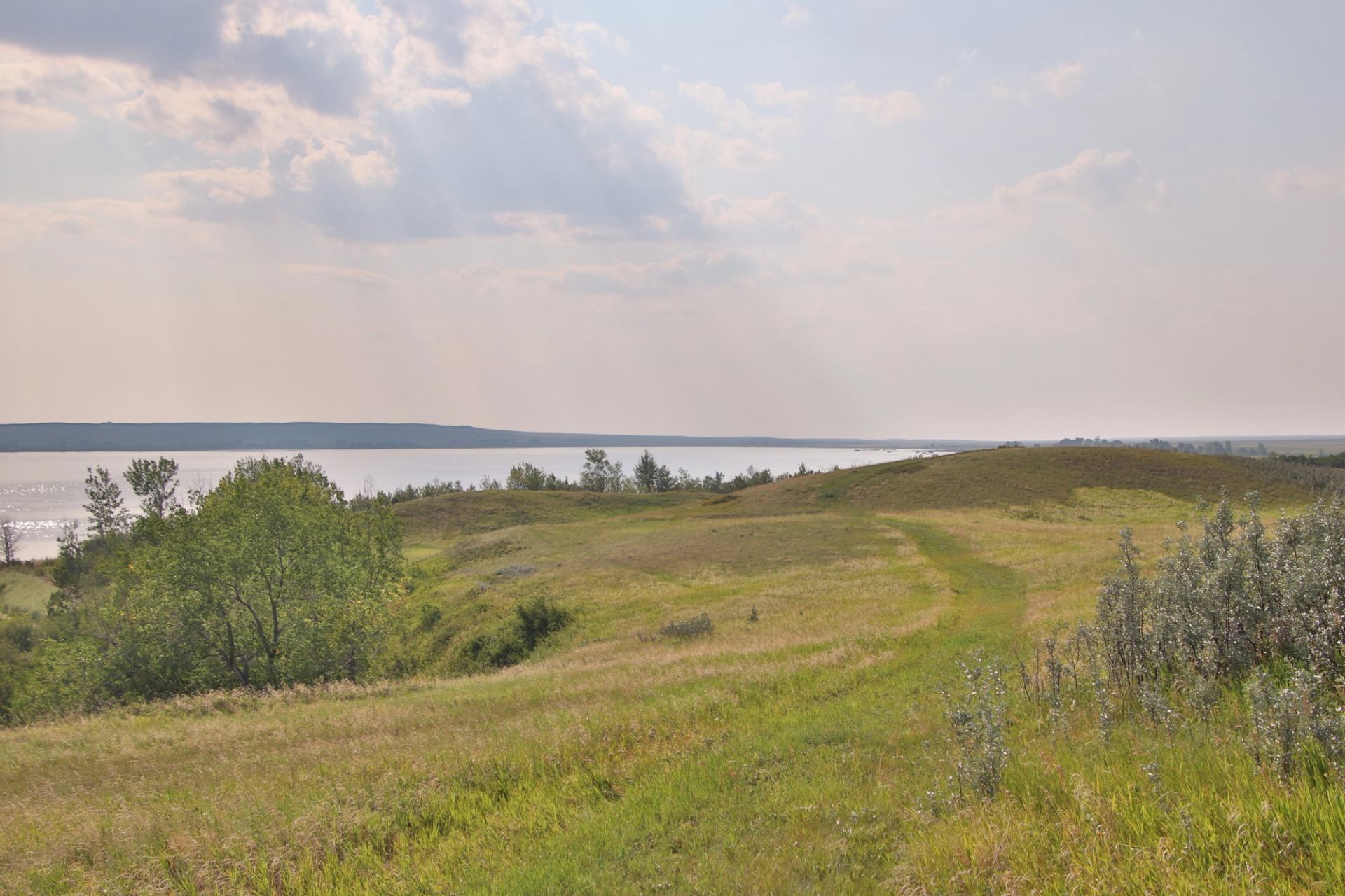 Grassy hills leading to a calm lake under hazy sunlight and scattered clouds.
