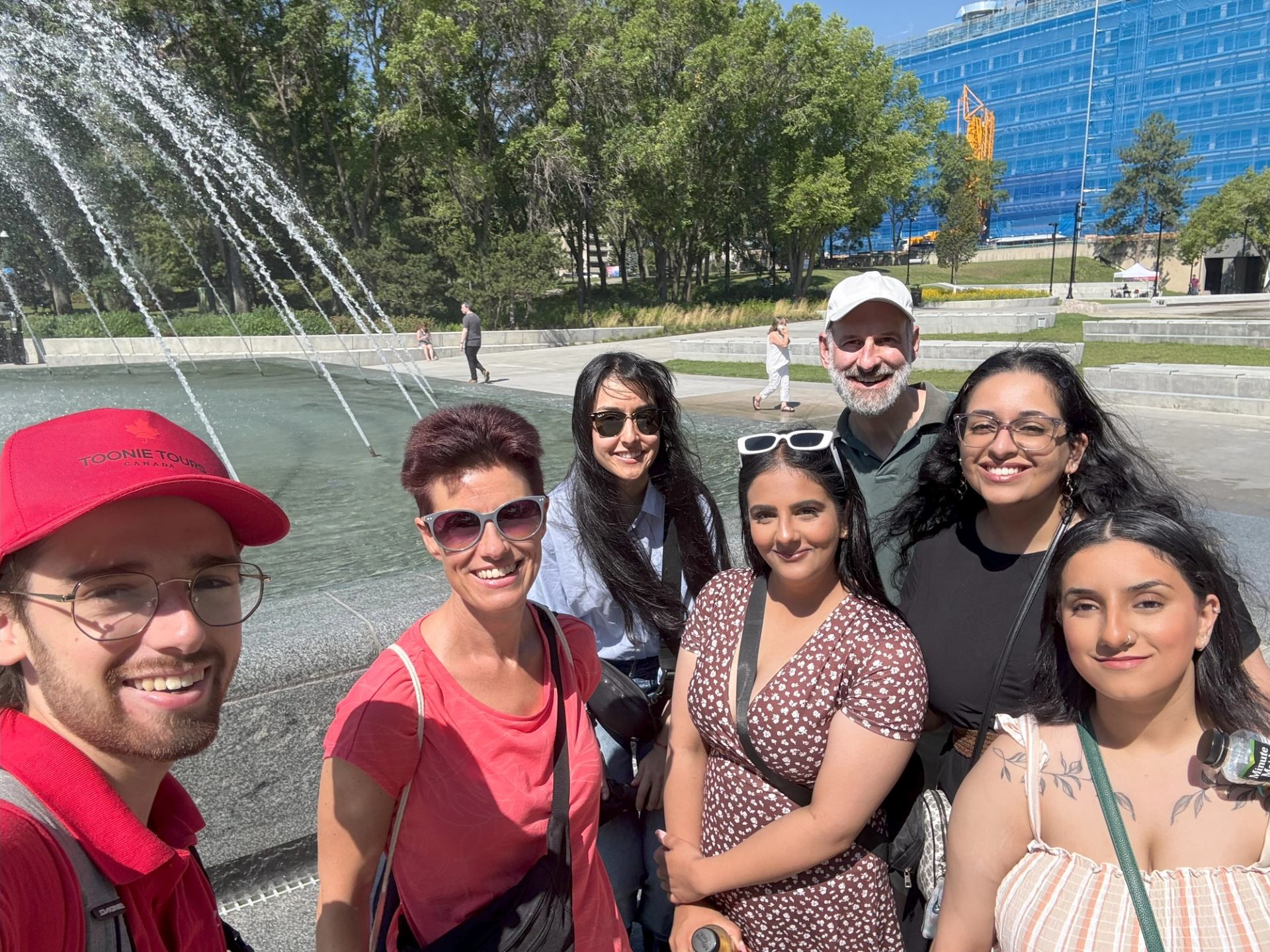 Seven people pose by a fountain with trees and a blue building in Edmonton.