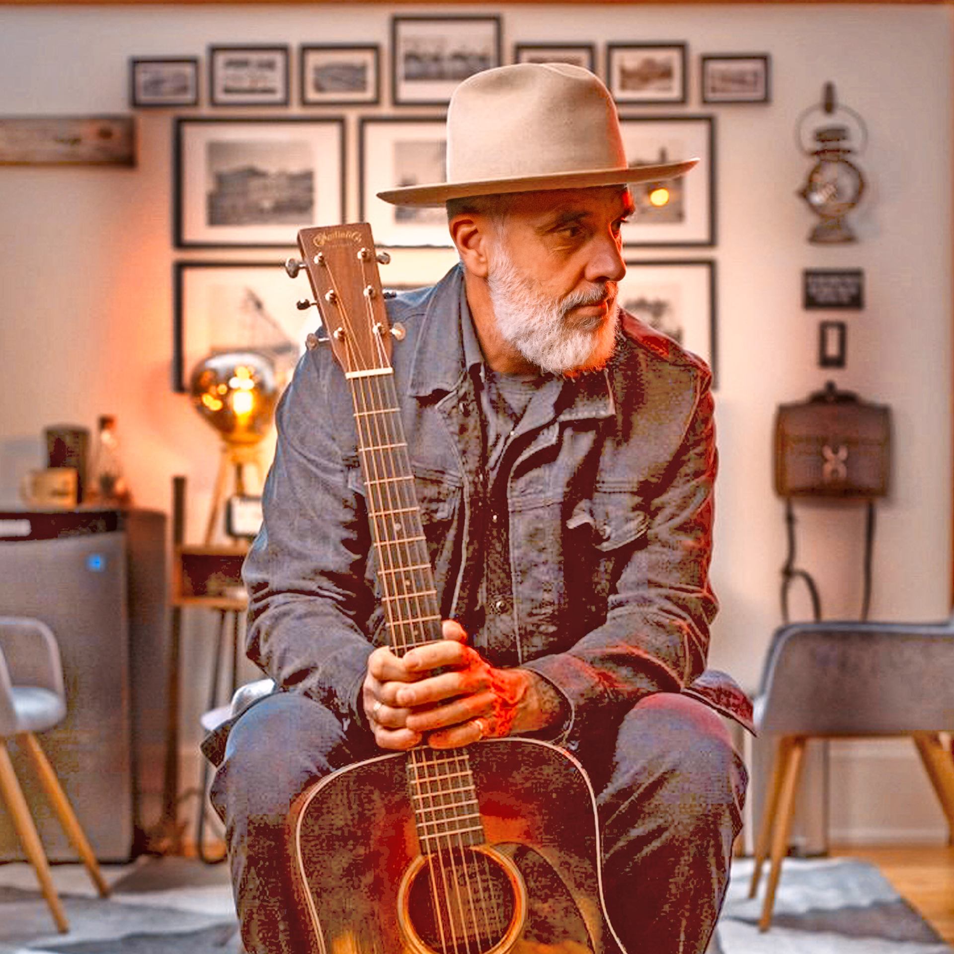 Bearded person in hat holding acoustic guitar, seated in warm, framed-photo filled room.