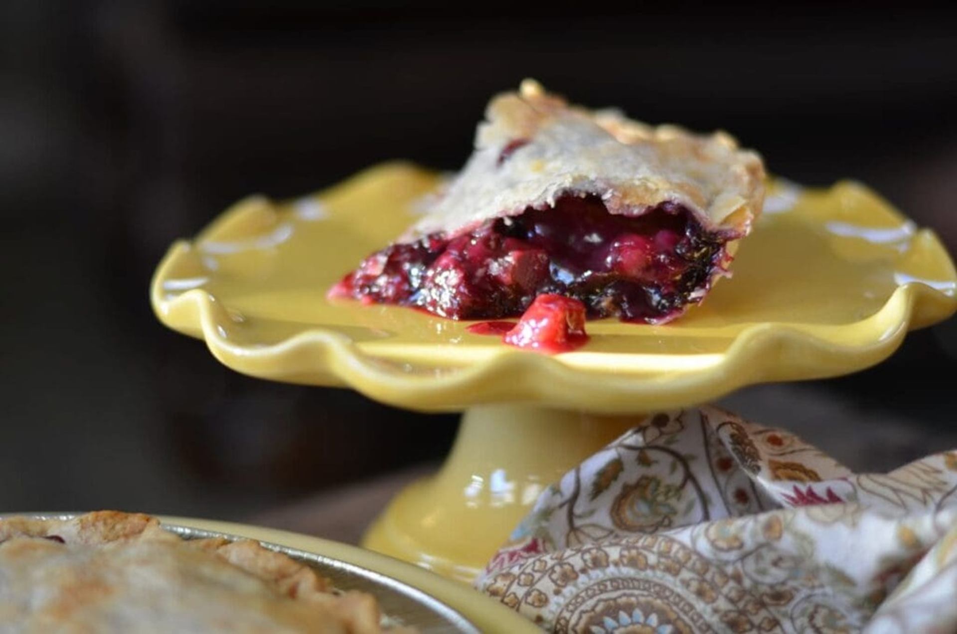 Slice of berry pie on yellow cake stand beside patterned cloth and whole pie.