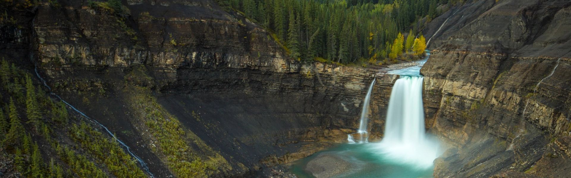 Wide waterfall plunging into turquoise pool between dark cliffs and dense evergreen forest.