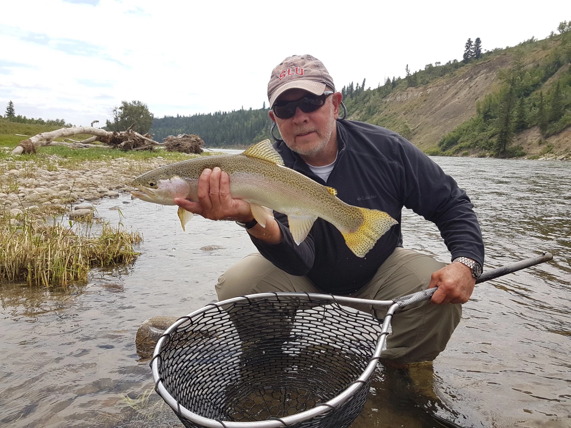 A man with sunglasses holds a fish and fishing net while squatting in a river.