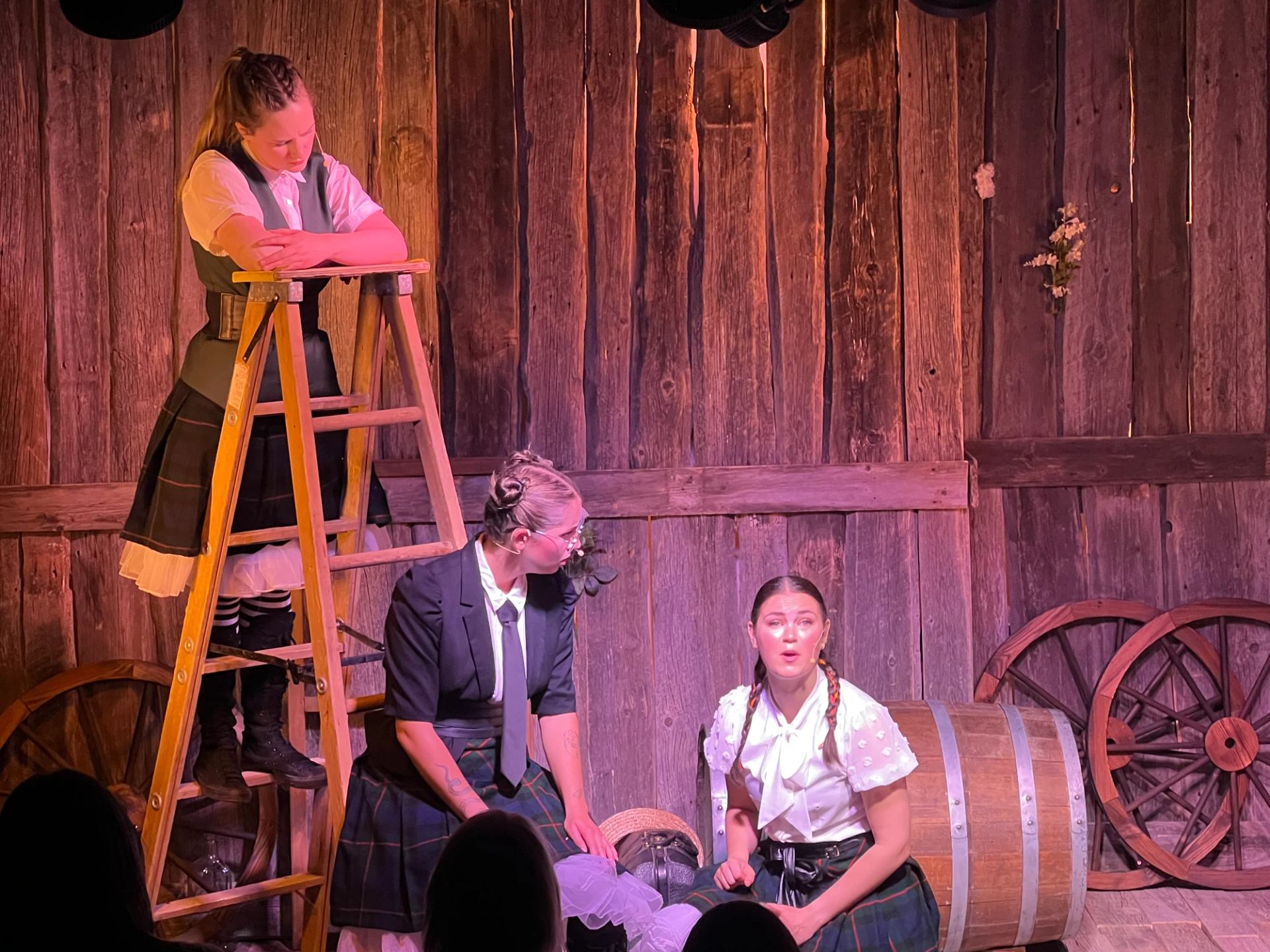 Three actors perform on rustic stage with ladder, barrel, and wagon wheels.