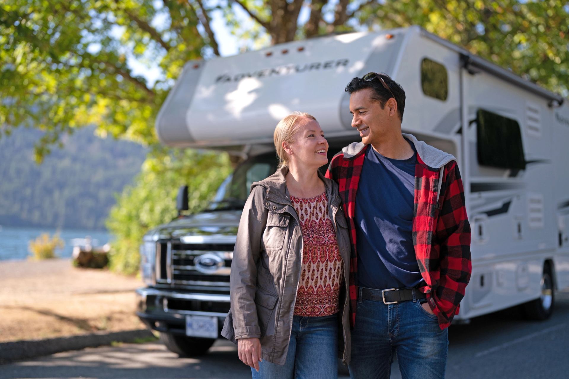 Couple walking near parked RV by trees and water in a scenic outdoor setting.