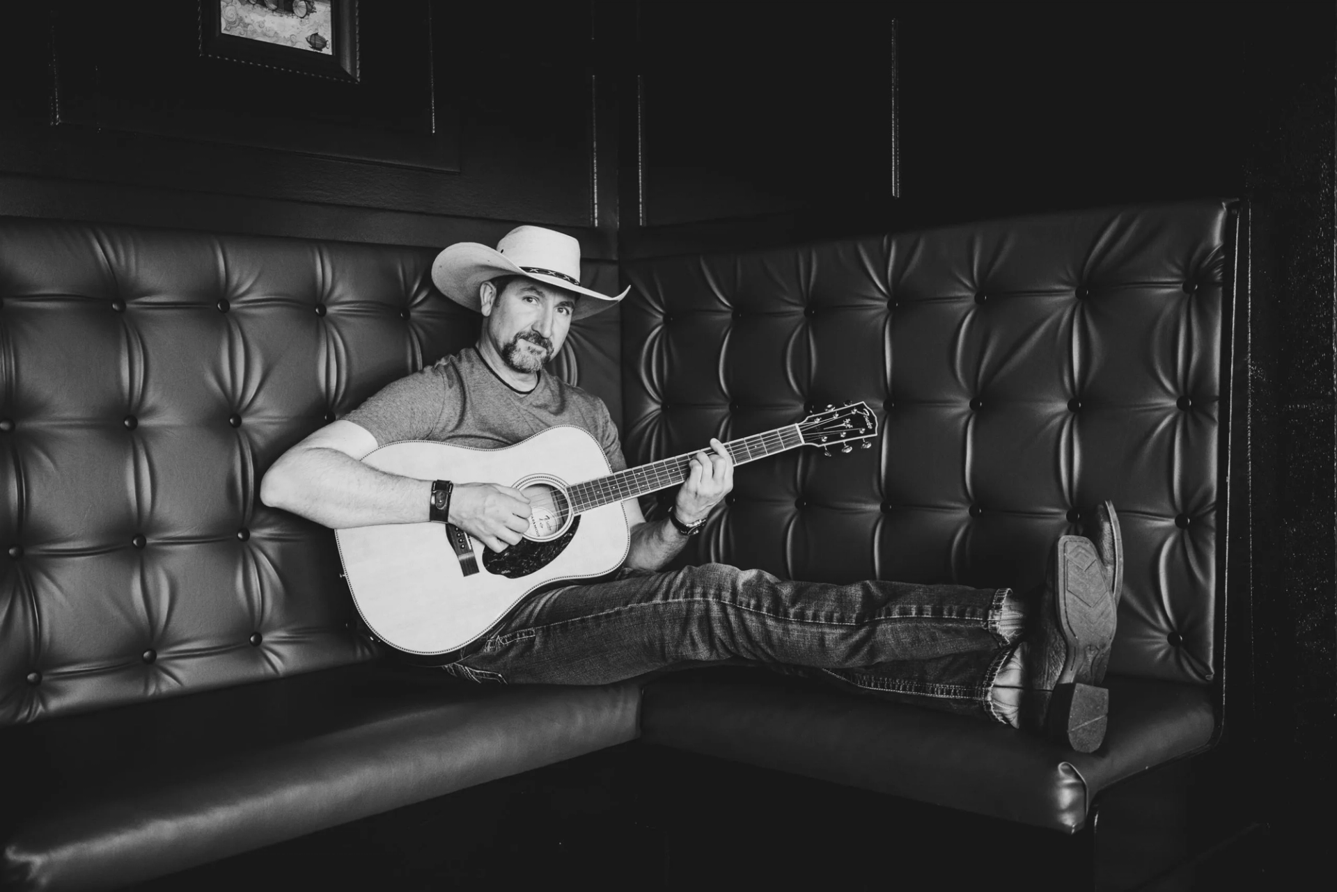 Man in cowboy hat plays acoustic guitar while reclining on tufted leather booth.