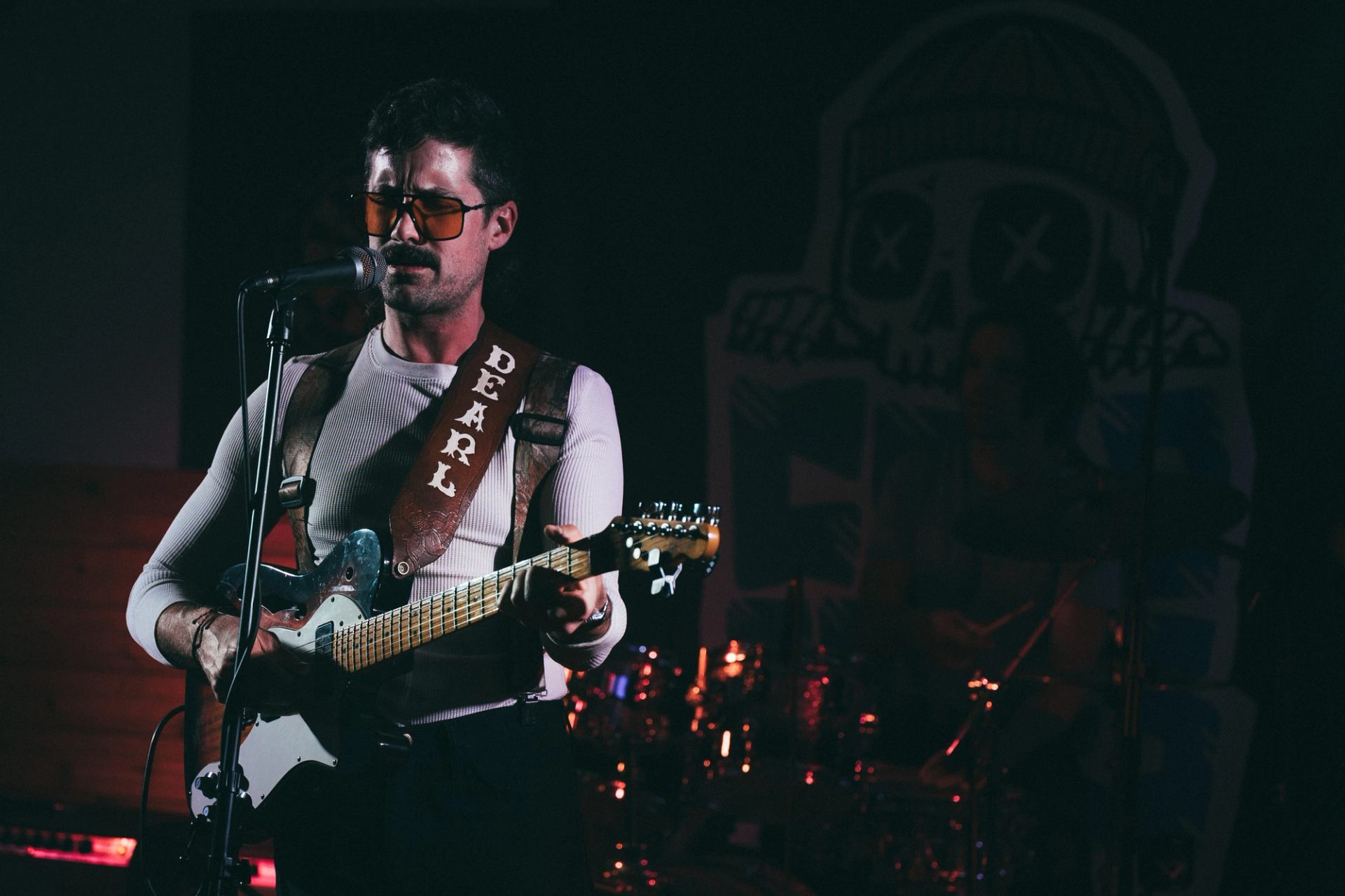 Performer playing electric guitar on stage with drum kit visible in the background