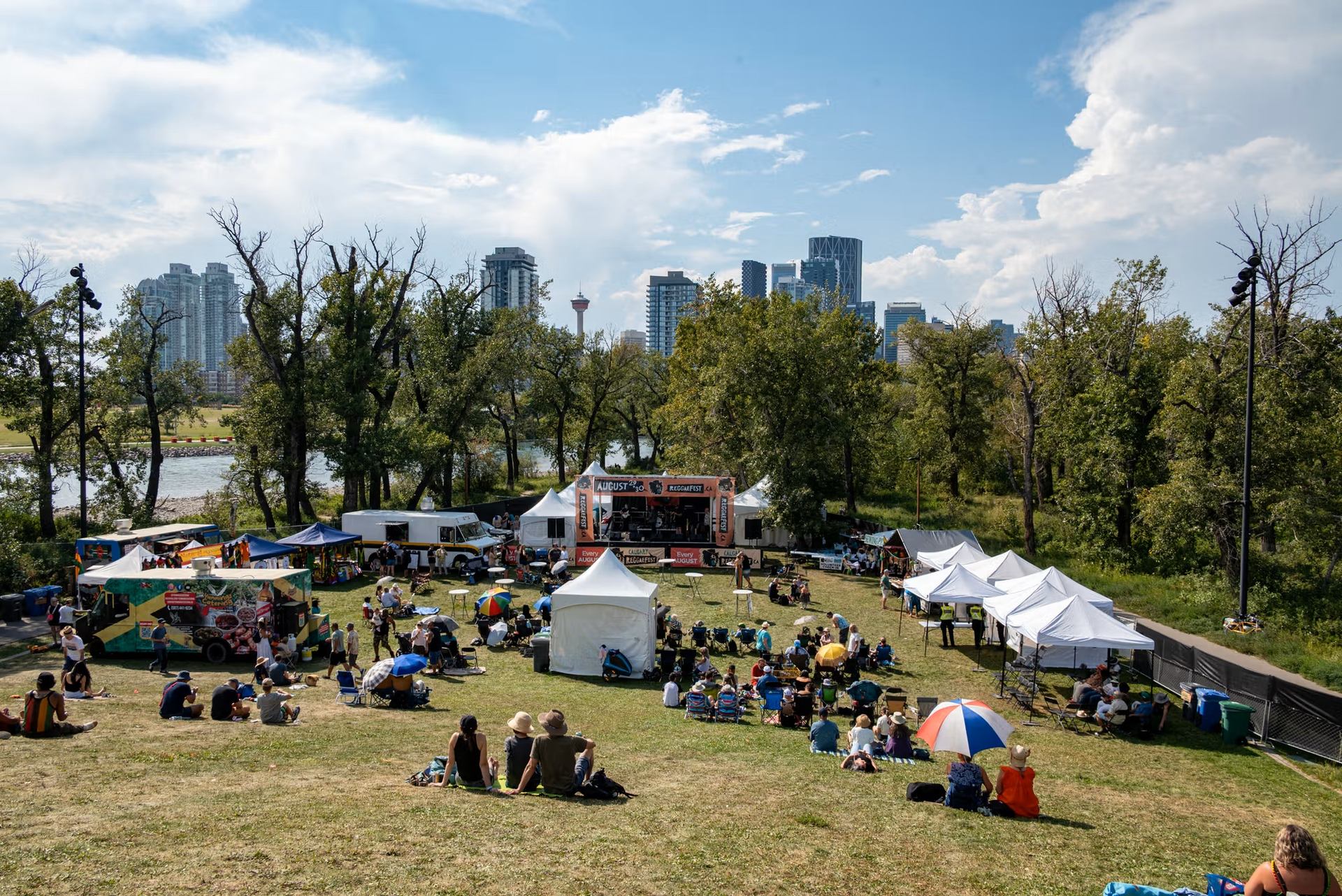Outdoor music festival crowd on grassy hill with stage, tents and city skyline backdrop.