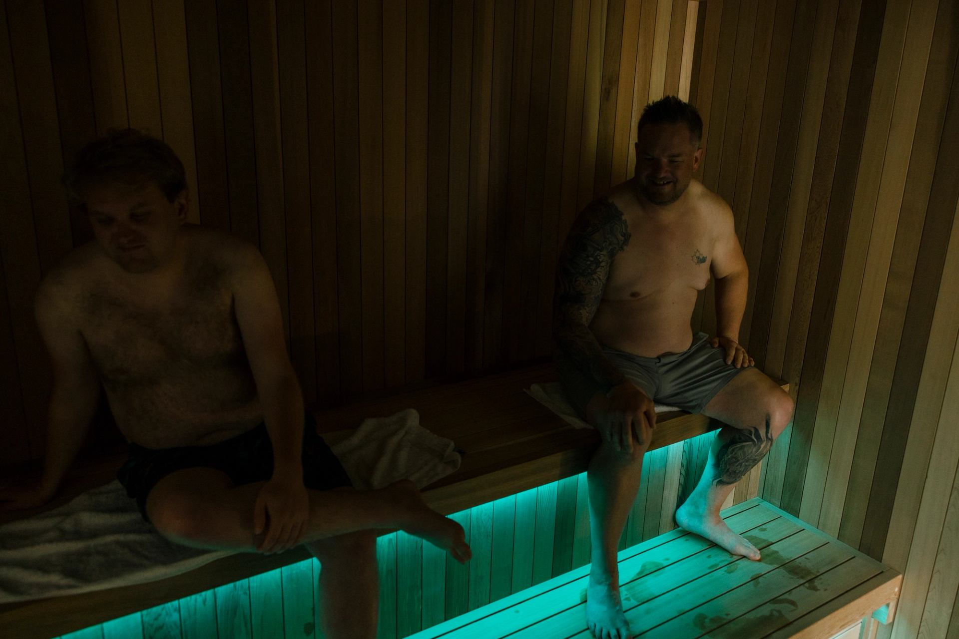 Two people sit on wooden sauna bench above teal floor lighting, wearing towels and shorts.