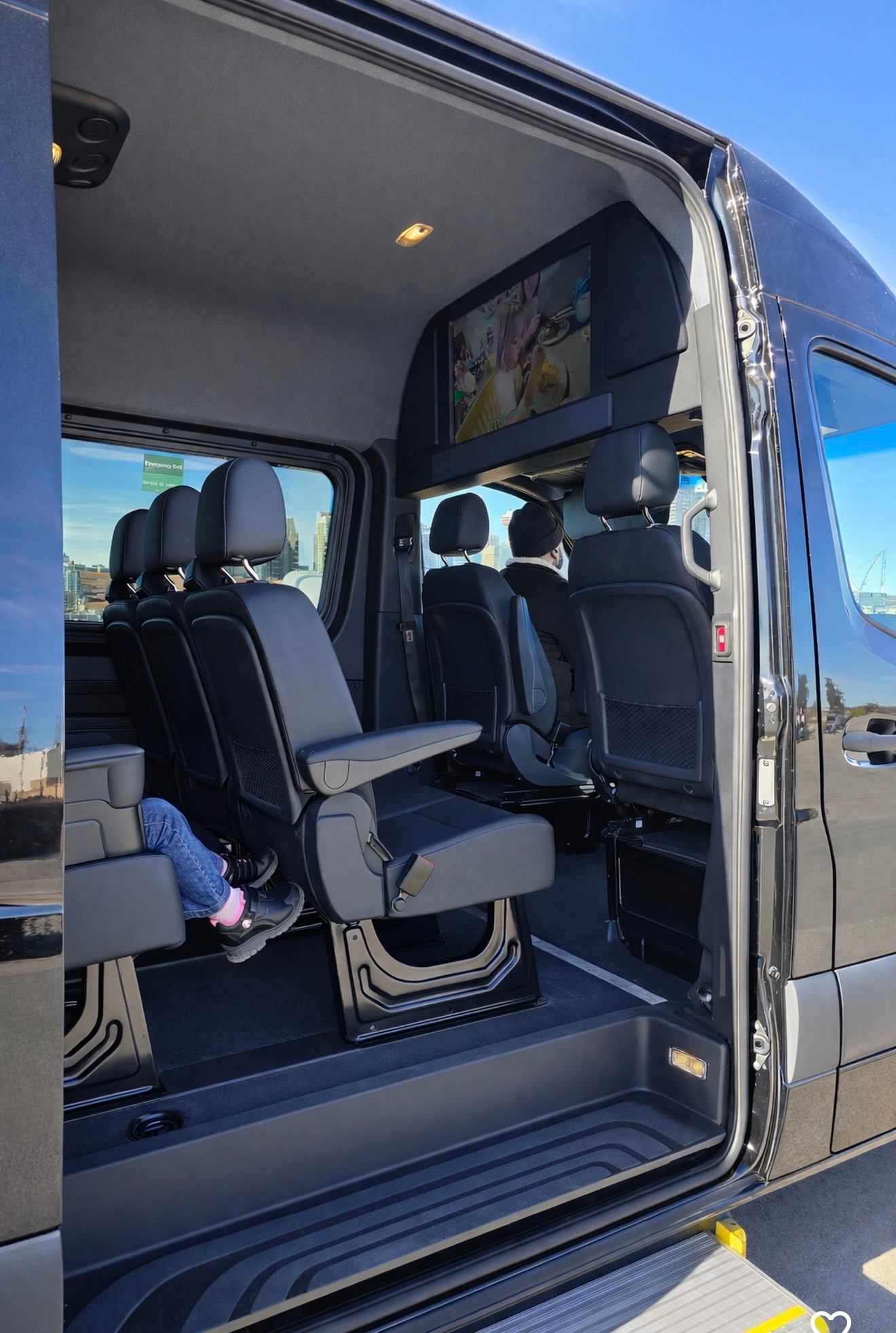 Interior of passenger van with black seats, ceiling screen and visible city skyline outside.