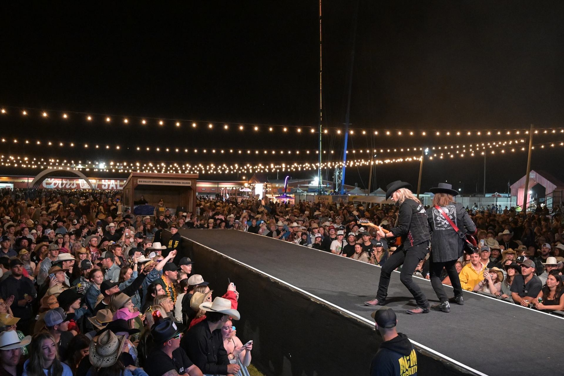 Country musicians perform on outdoor stage above large crowd under string lights at night.