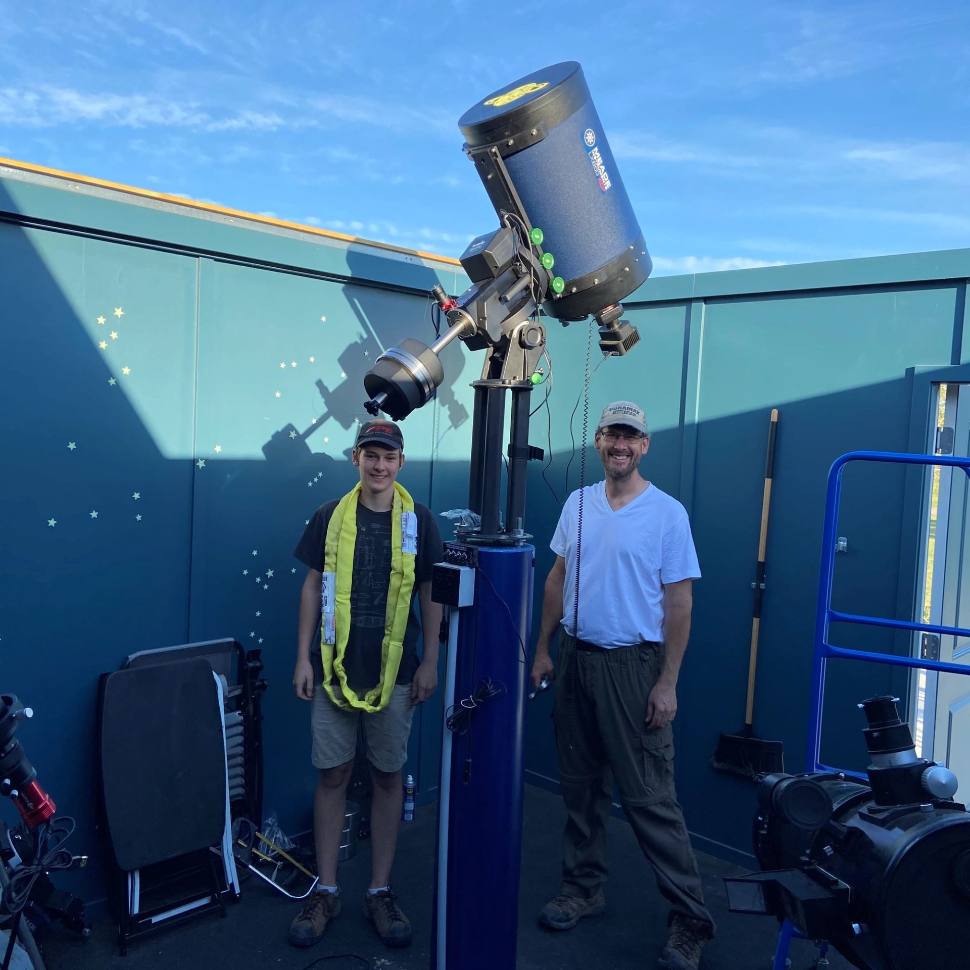 Two people beside large telescope at outdoor observatory with blue walls.