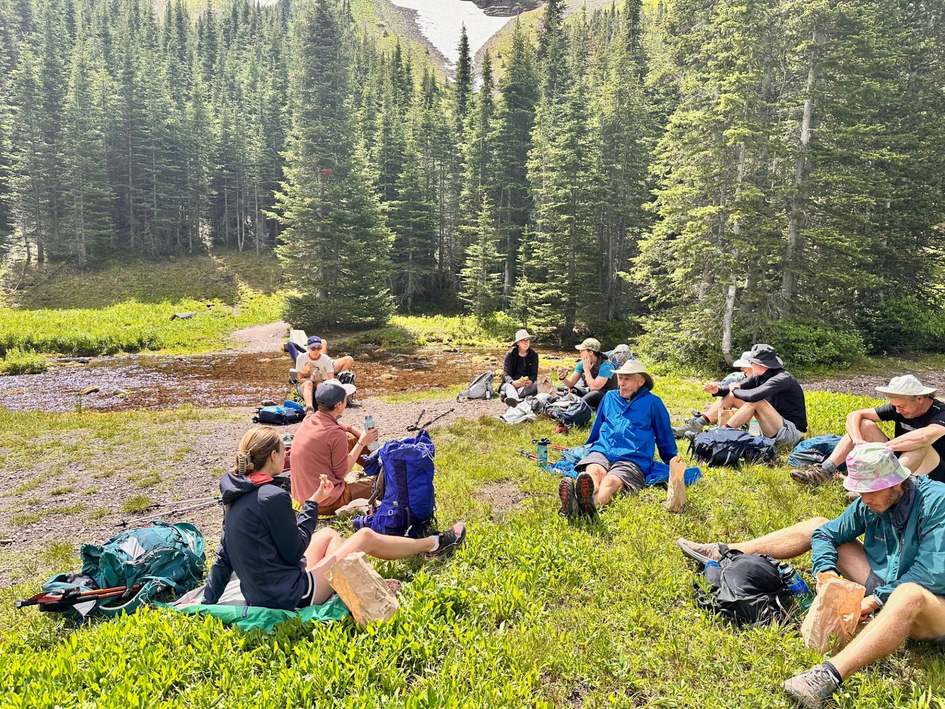 Hikers sit on grass with backpacks, eating lunch beside a small forest pond.