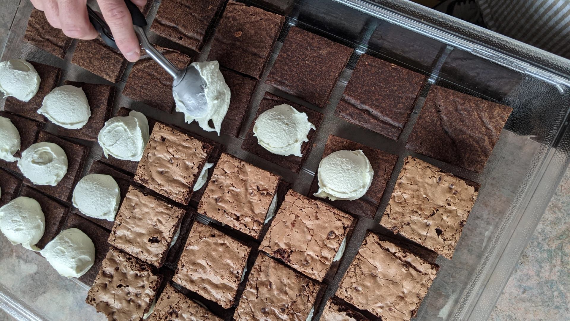 Brownies being topped with a scoop of whipped cream. The brownies are arranged in neat squares in a container.