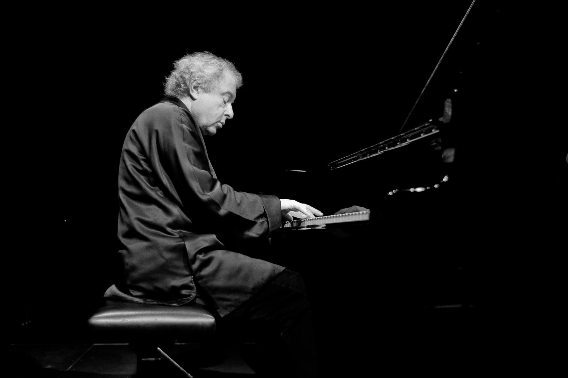 Pianist in dark clothing plays grand piano onstage against a black background.