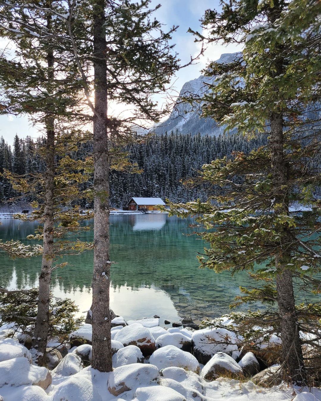 A serene lake with clear, turquoise water is framed by snow-dusted trees. A cozy cabin is nestled at the forest's edge, with mountains in the background.