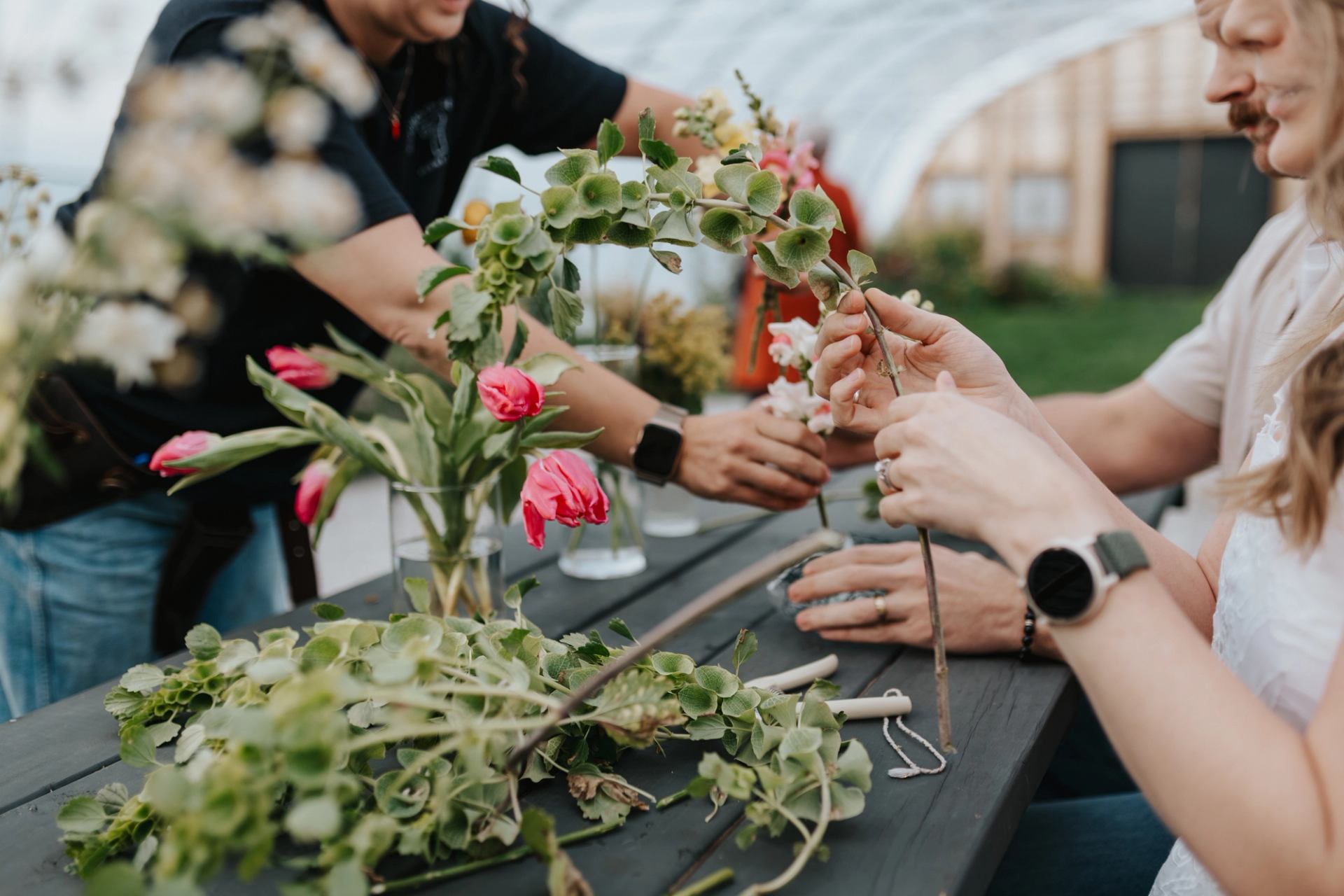 Hands working with fresh flowers and greenery at a table inside a greenhouse.