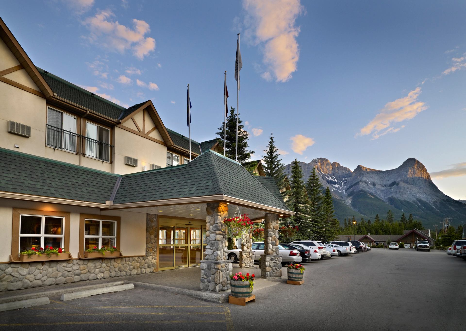 Hotel with stone facade, flags, and mountain views surrounded by pine trees.