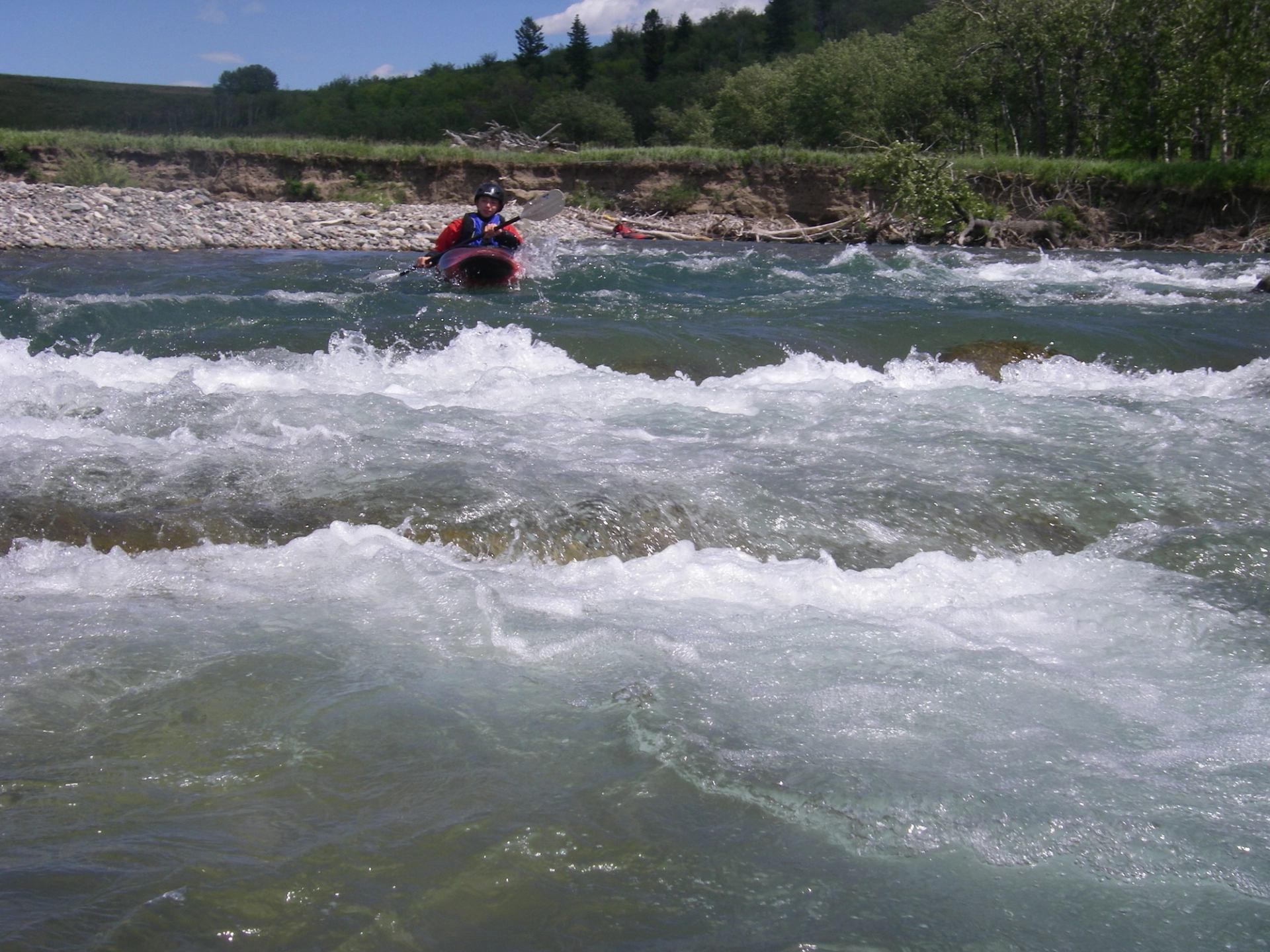 Kayaker approaching small river rapids surrounded by greenery.