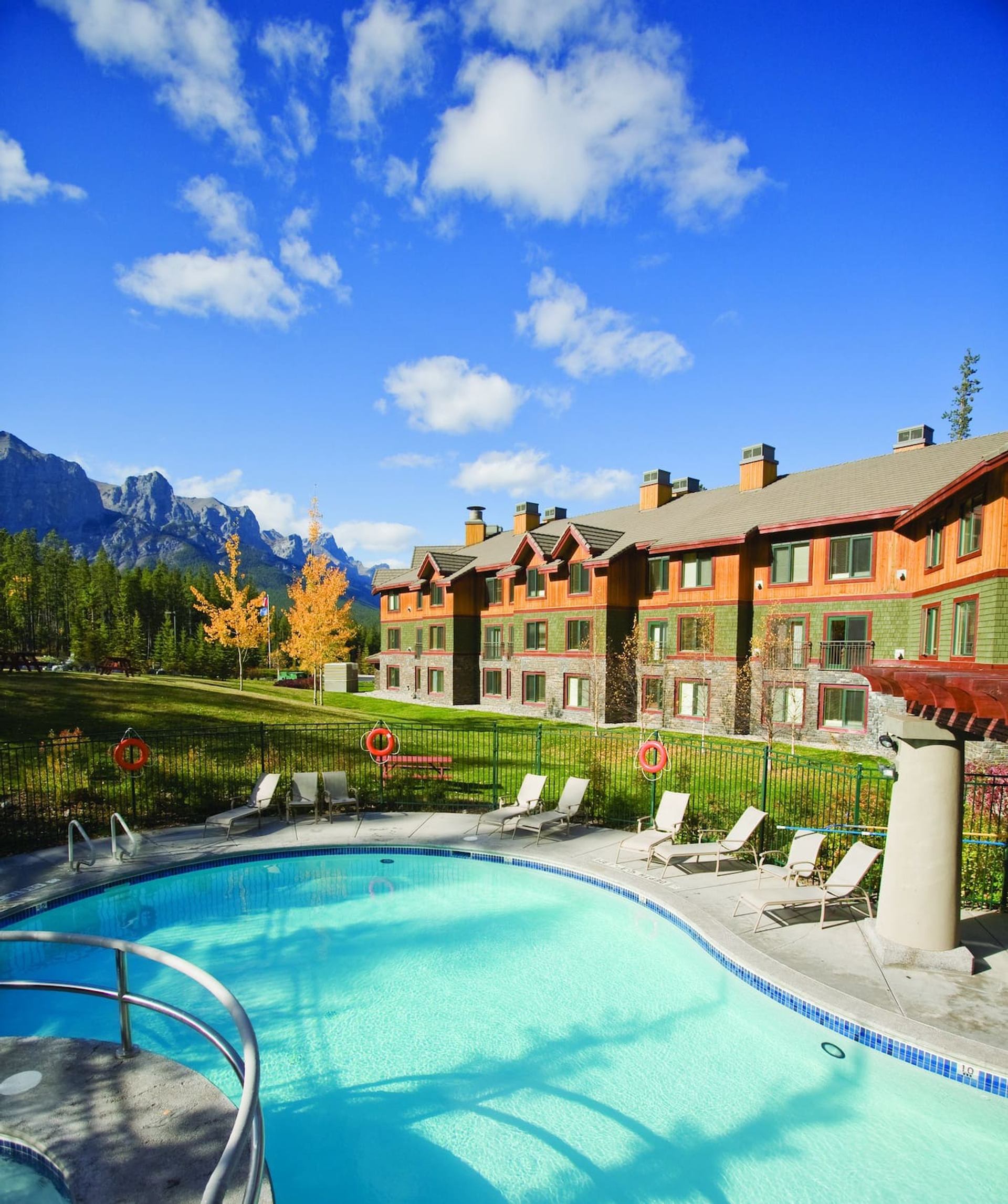 Outdoor pool with lounge chairs, mountain backdrop, and resort-style buildings.