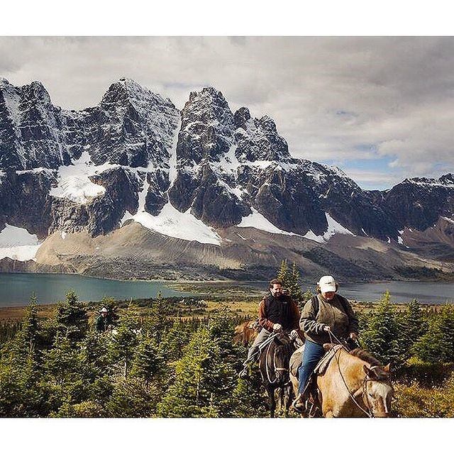 Two horseback riders ride towards the viewer with mountains and a lake in the background.
