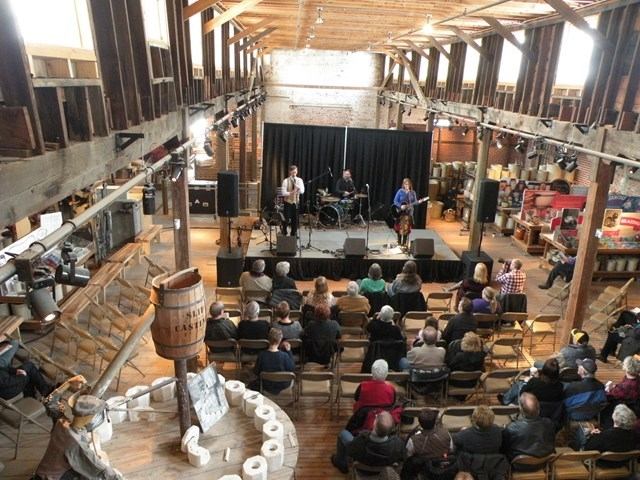 Audience seated in a wooden hall watching a band perform on stage at Tongue on the Post Festival.
