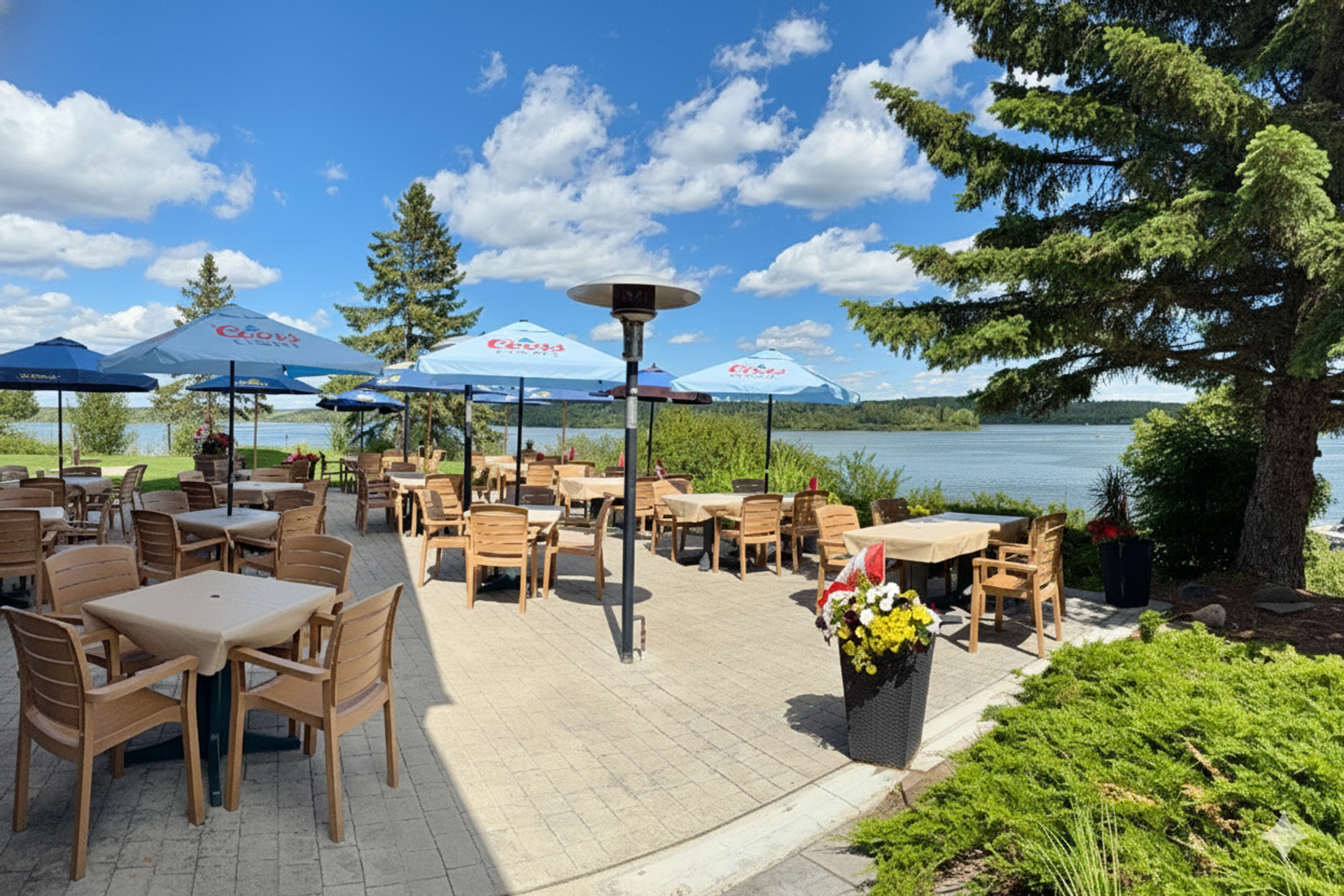 Restaurant patio with wooden tables, blue umbrellas and lake view under scattered clouds.