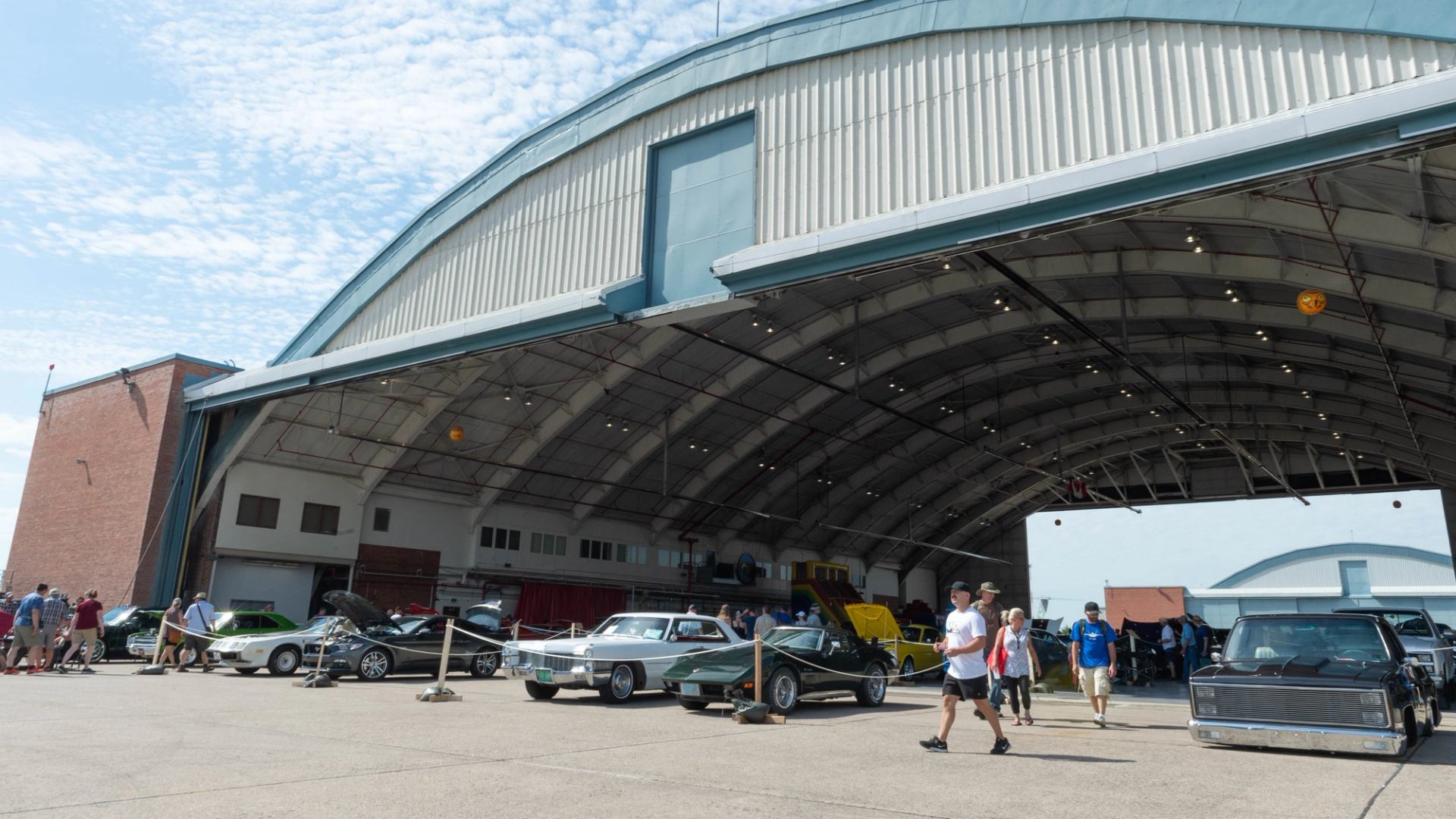 People and cars gathered around large open hangar under partly cloudy sky.