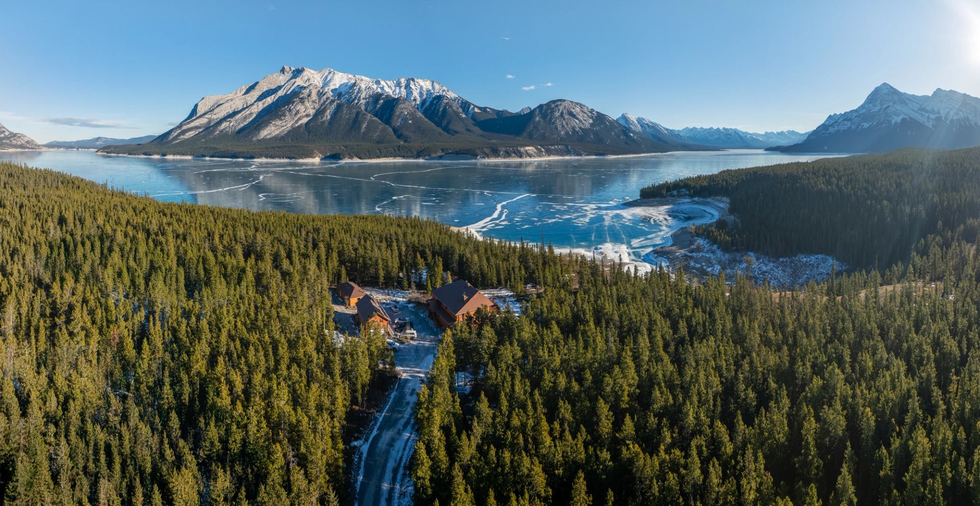 Aerial view of a lakeside lodge surrounded by a pine forest, with a partially frozen lake and snow-capped mountains.