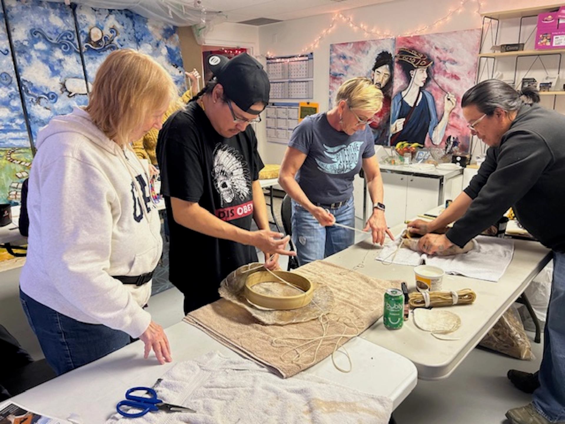 People stand around tables, crafting circular items with string in an art studio.