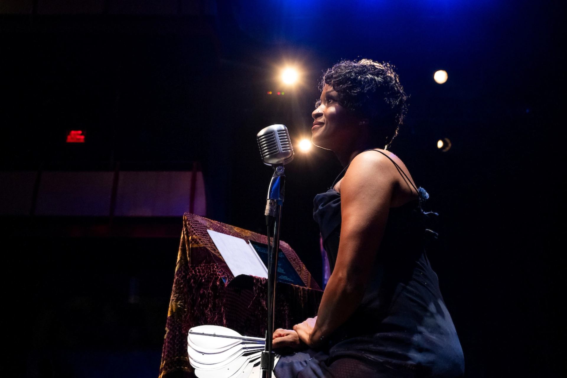 A woman with dark curly hair sings into a vintage microphone on a dark stage under spotlights.