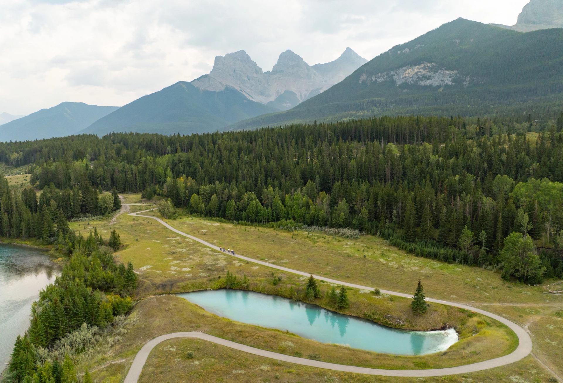 Mountain valley with a winding road, turquoise pond, and dense evergreen forest under cloudy sky.